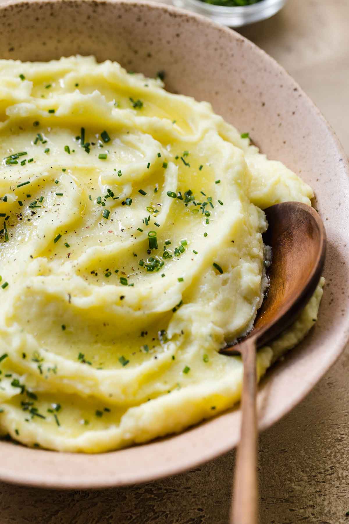 Close up photo of mashed potatoes in a serving bowl, sprinkled with minced chives, and a wooden spoon resting in the bowl.