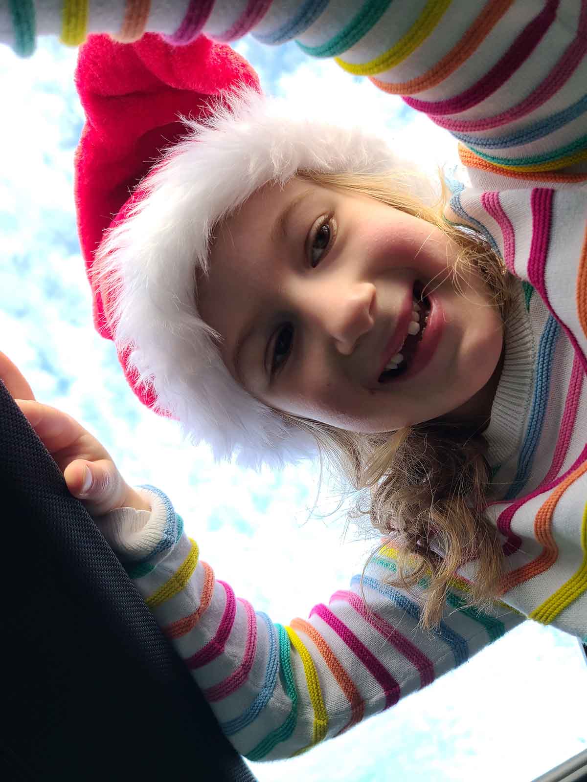 Little girl in santa hat and striped sweater standing in moon roof opening with blue sky in background.