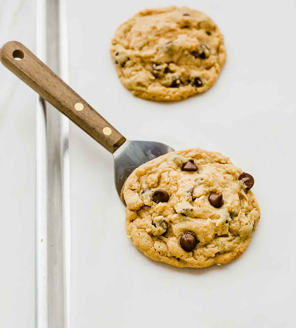 Stainless steel spatula slid underneath of a peanut butter oatmeal chocolate chip cookie on a parchment-lined baking sheet.