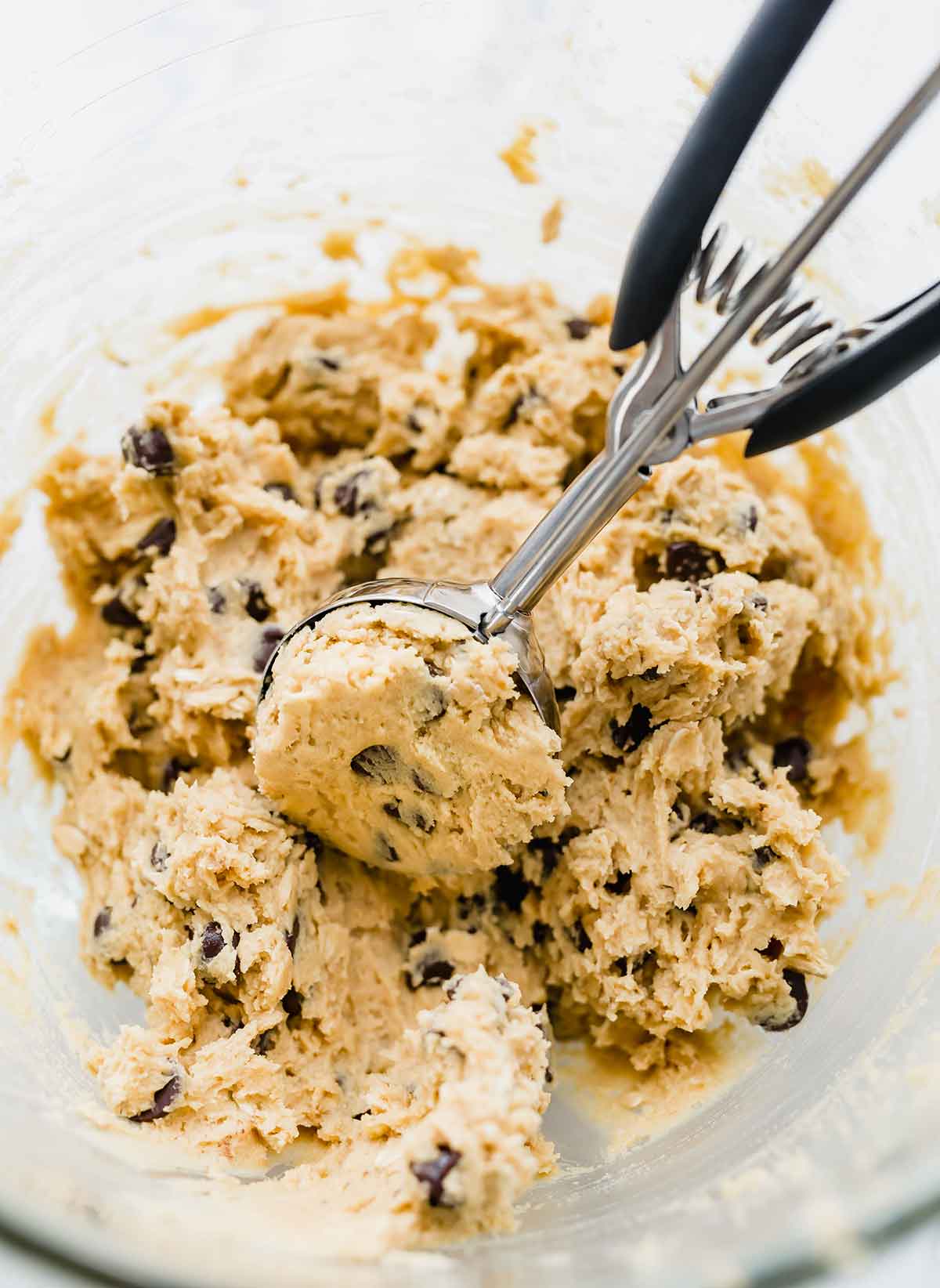 A cookie scoop sccoping cookie dough for peanut butter oatmeal chocolate chip cookies out of a glass bowl.