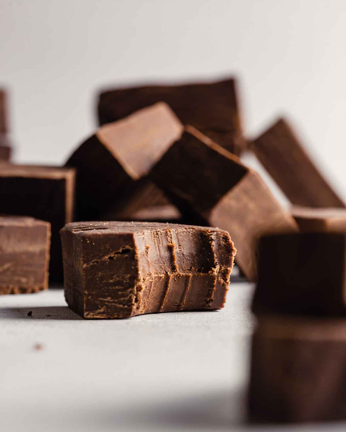 A single piece of fudge with a bite taken out of it in the foreground with squares of fudge stacked in the background.
