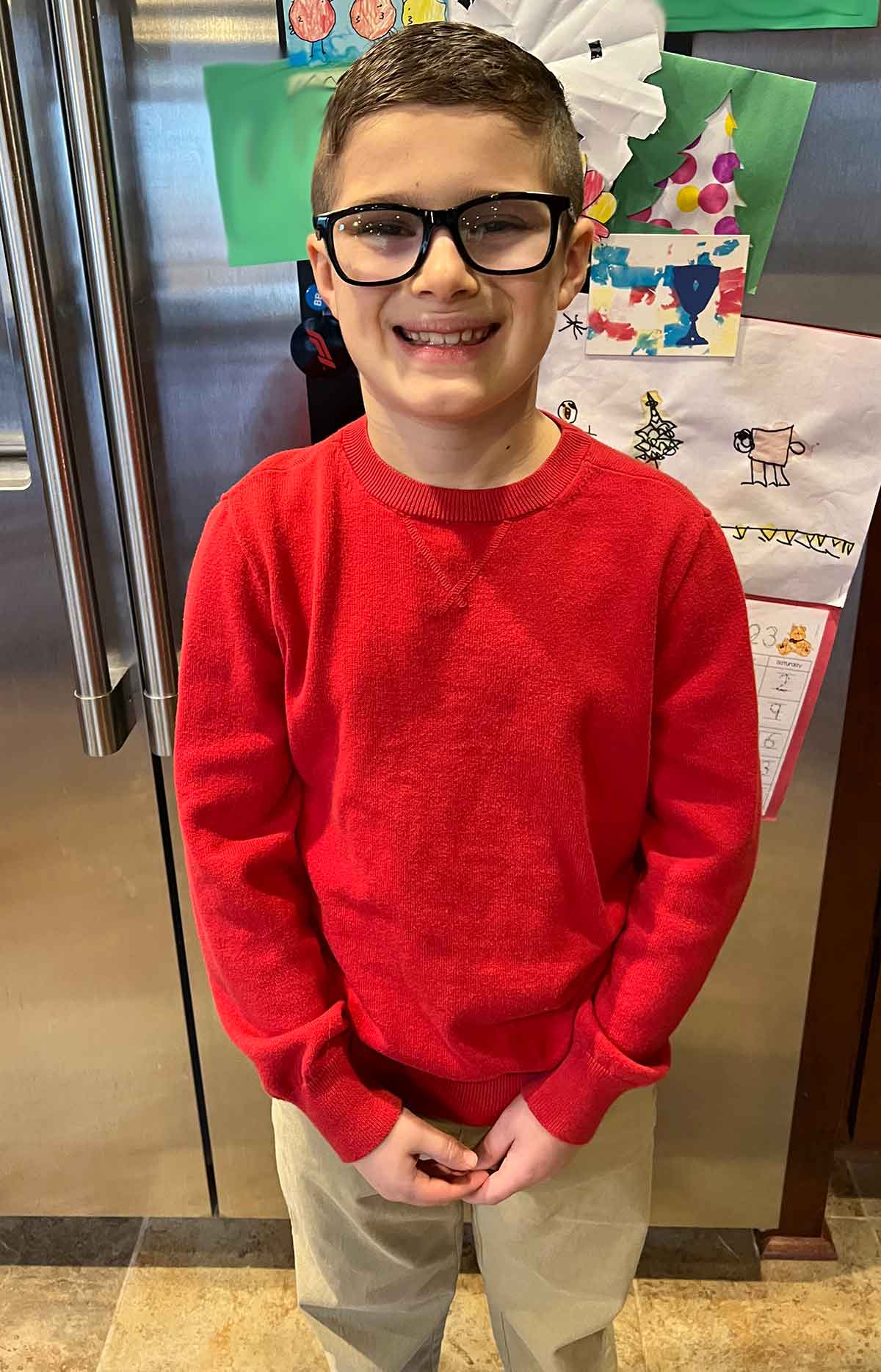 Little boy in khaki pants and red sweater smiling in front of a refrigerator.