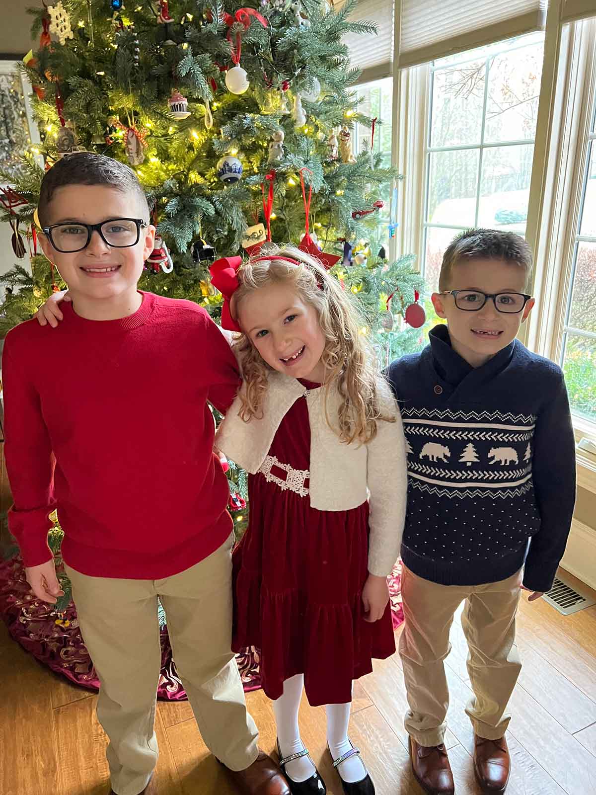 Two boys and a girl standing in front of a lit Christmas tree wearing Christmas attire.