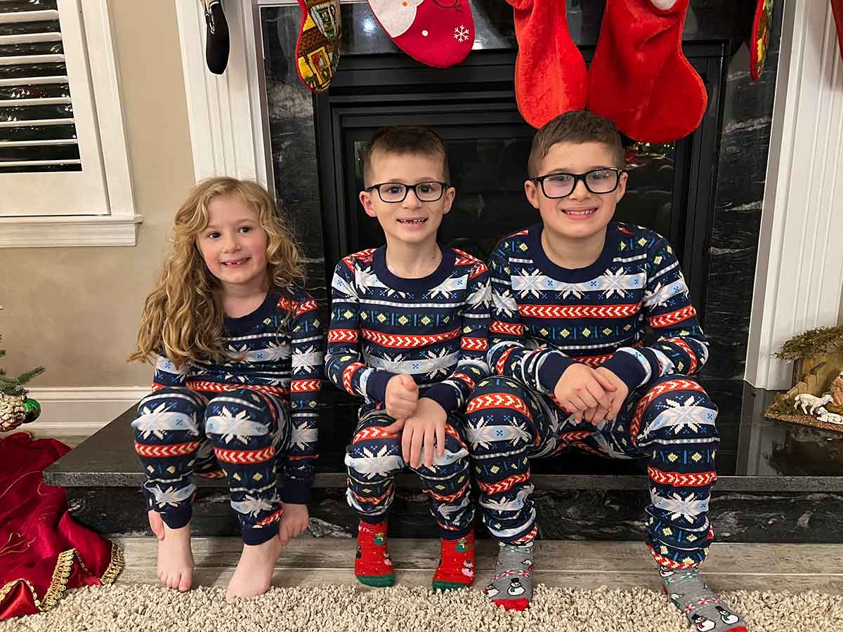 Three children in Christmas pajamas sitting on the hearth of a fireplace.