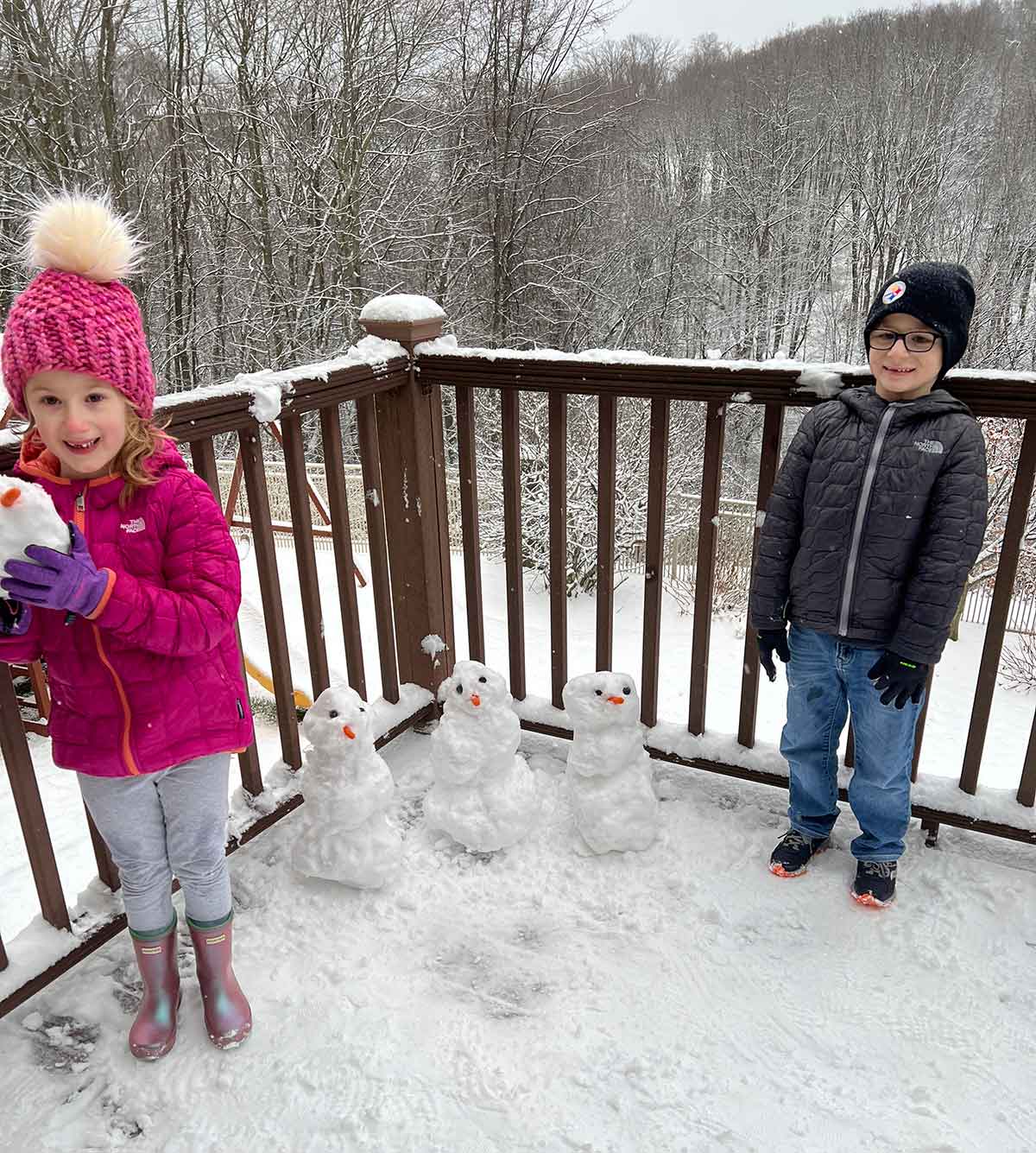 A girl and boy standing on a snowy deck with three small snowmen between them.