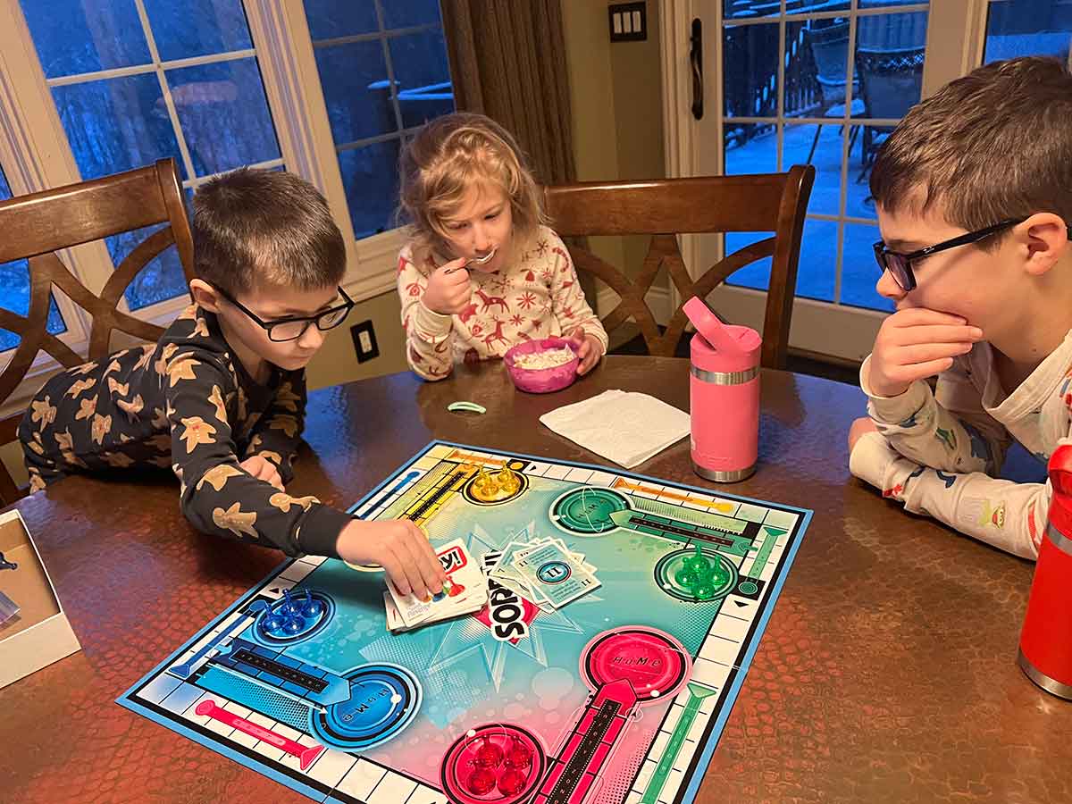 Three kids sitting around the board game Sorry at a kitchen table.