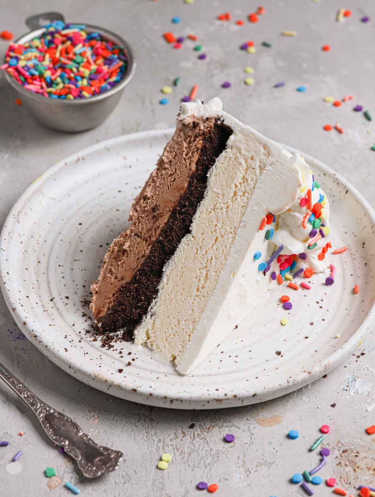 A slice of ice cream cake on a plate with layers of chocolate ice cream, fudge, Oreos, vanilla ice cream, and whipped cream.