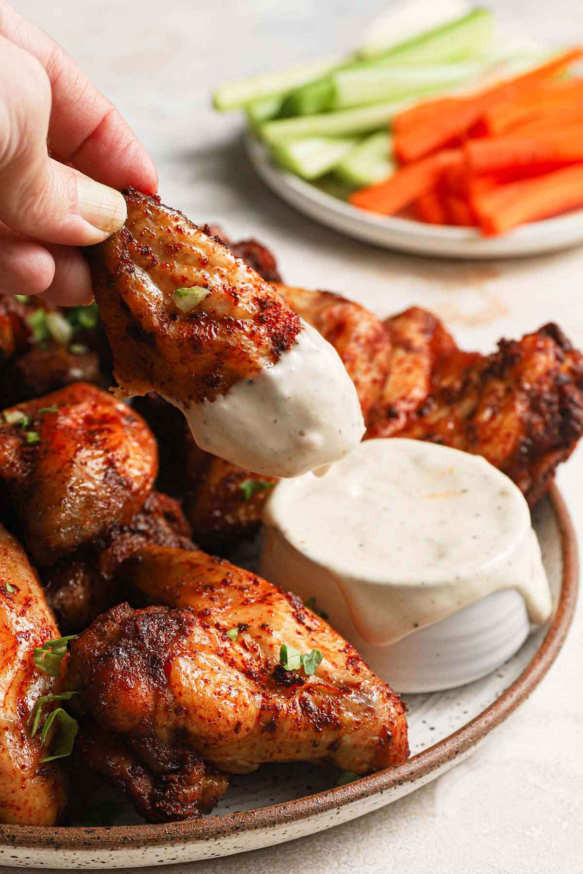 Platter of chicken wings with one being dipped into ranch dressing, and a plate of carrots and celery sticks in the background.