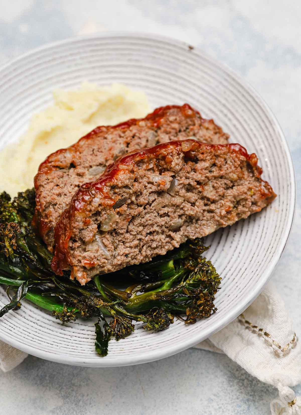 Two slices of meatloaf on a plate atop a pile of mashed potatoes and roasted broccolini.
