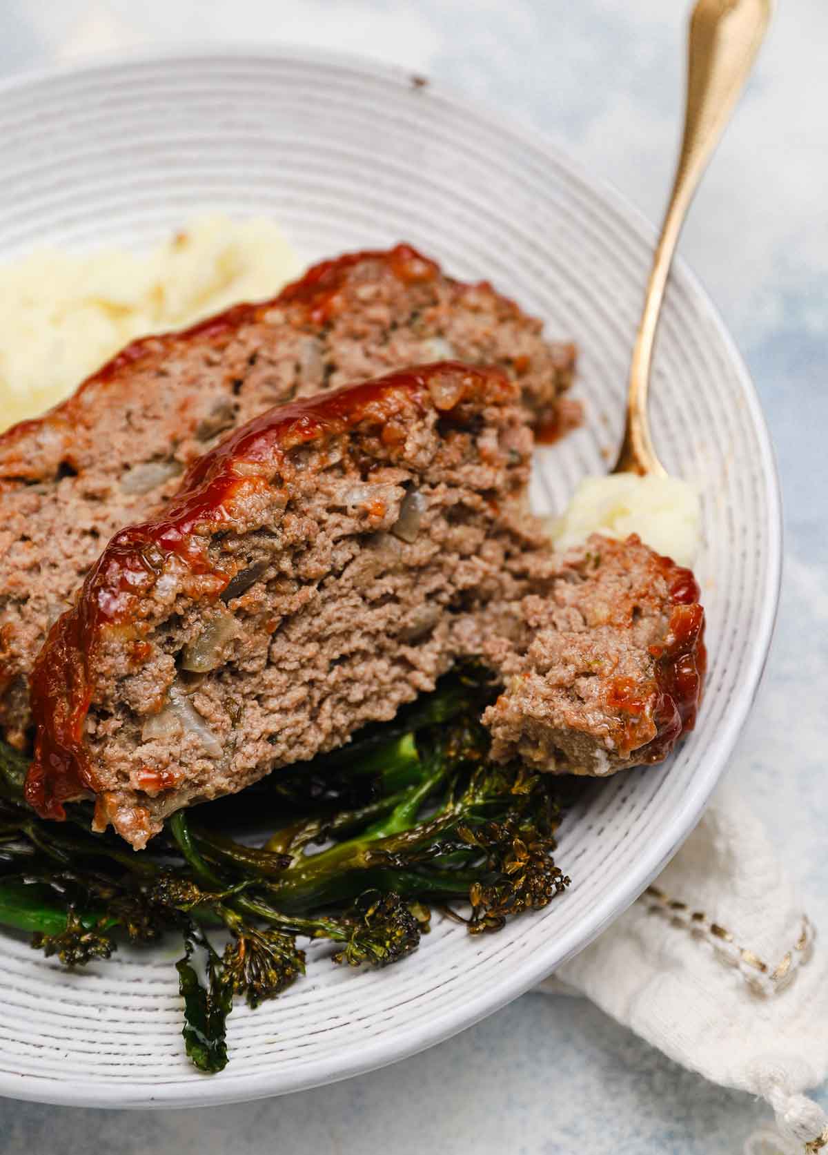 Fork cutting away a bite of meatloaf on a plate where there's another slice of meatloaf, mashed potatoes, and greens.