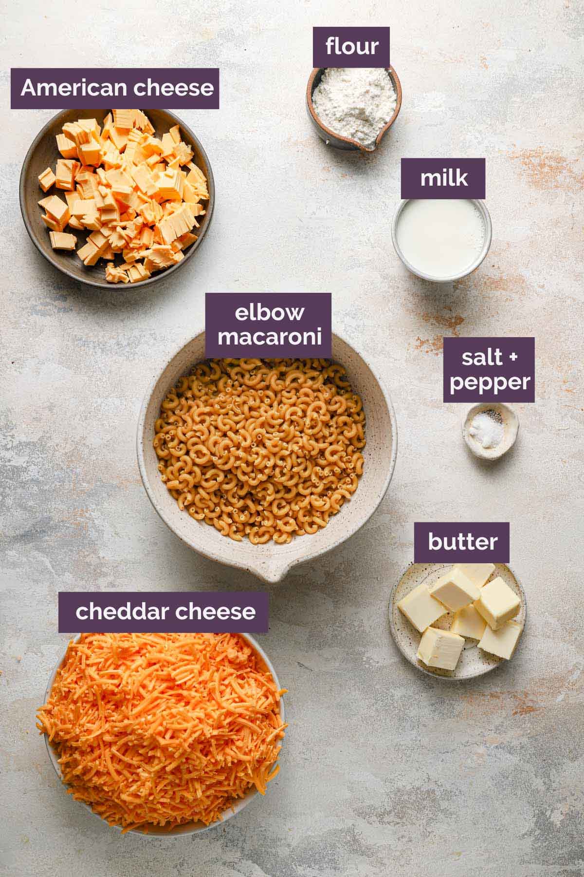 Ingredients for stovetop mac and cheese prepped and labeled: American cheese, flour, milk, elbow macaroni, salt, pepper, butter, and cheddar cheese.