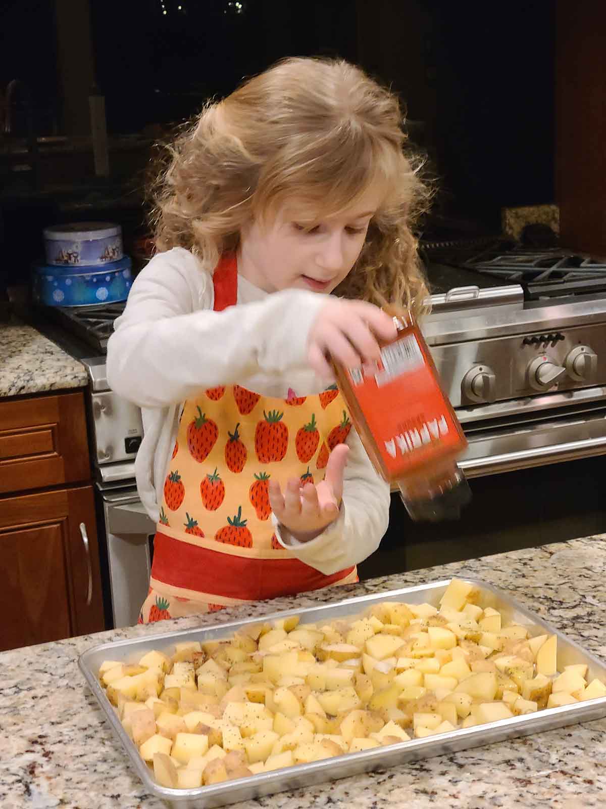 Little girl in strawberry apron seasoning potatoes with paprika.
