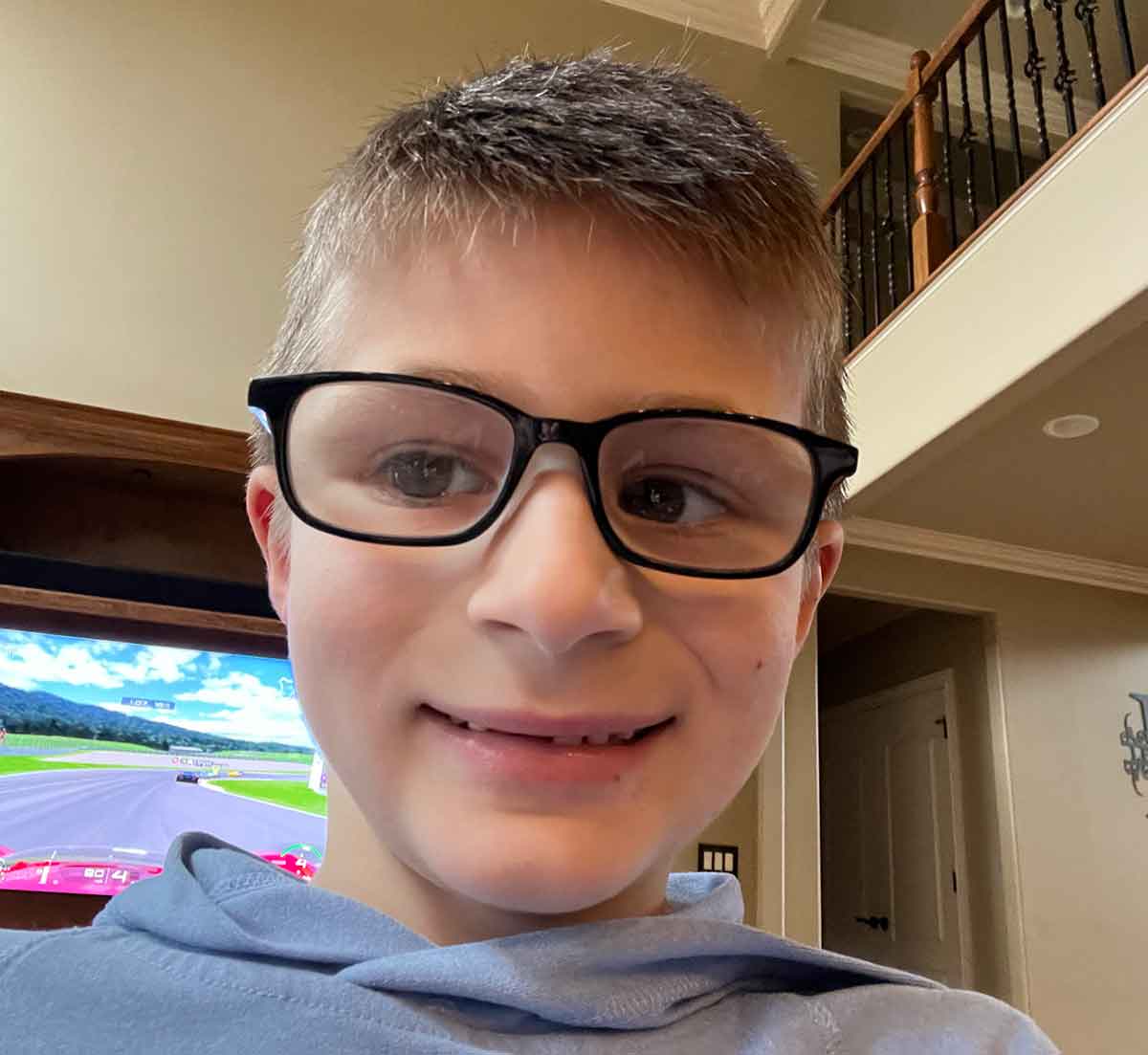Little boy in black glasses taking a selfie with a TV and upstairs railing in the background.