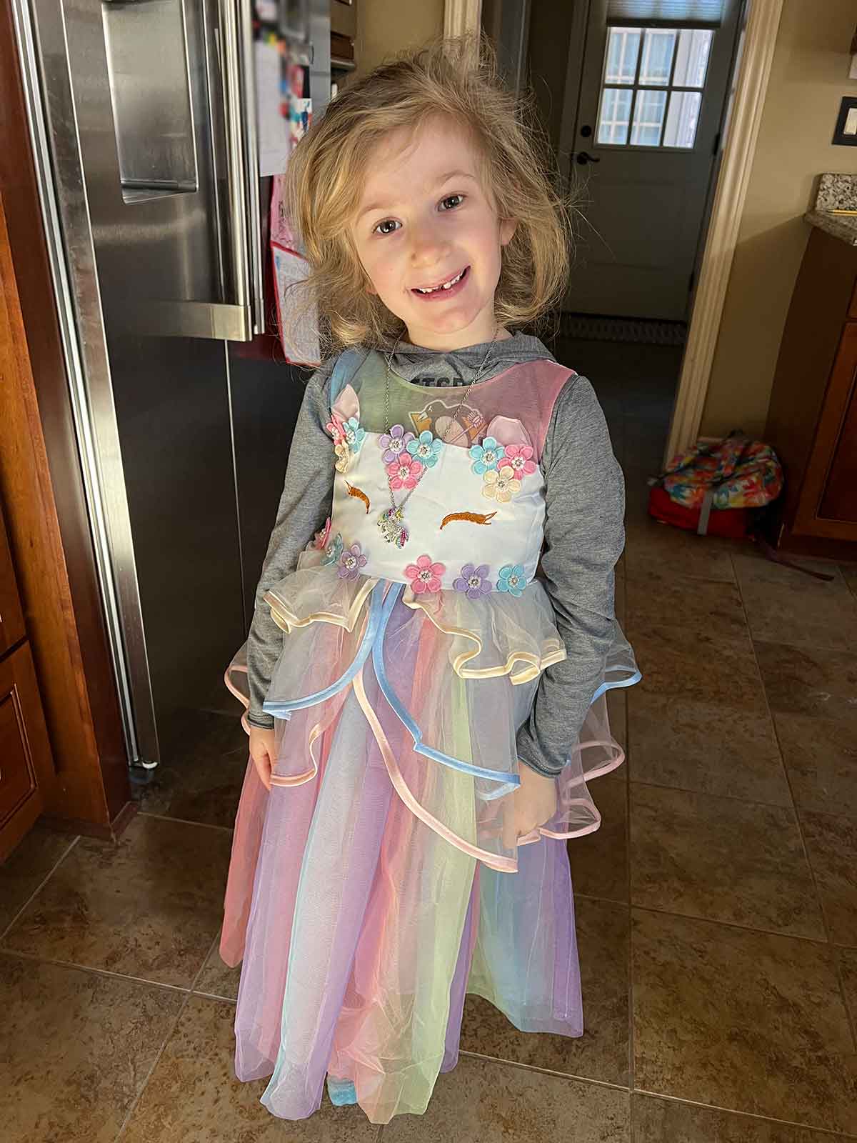 Little girl in a unicorn tulle dress standing in a kitchen with a refrigerator behind her.