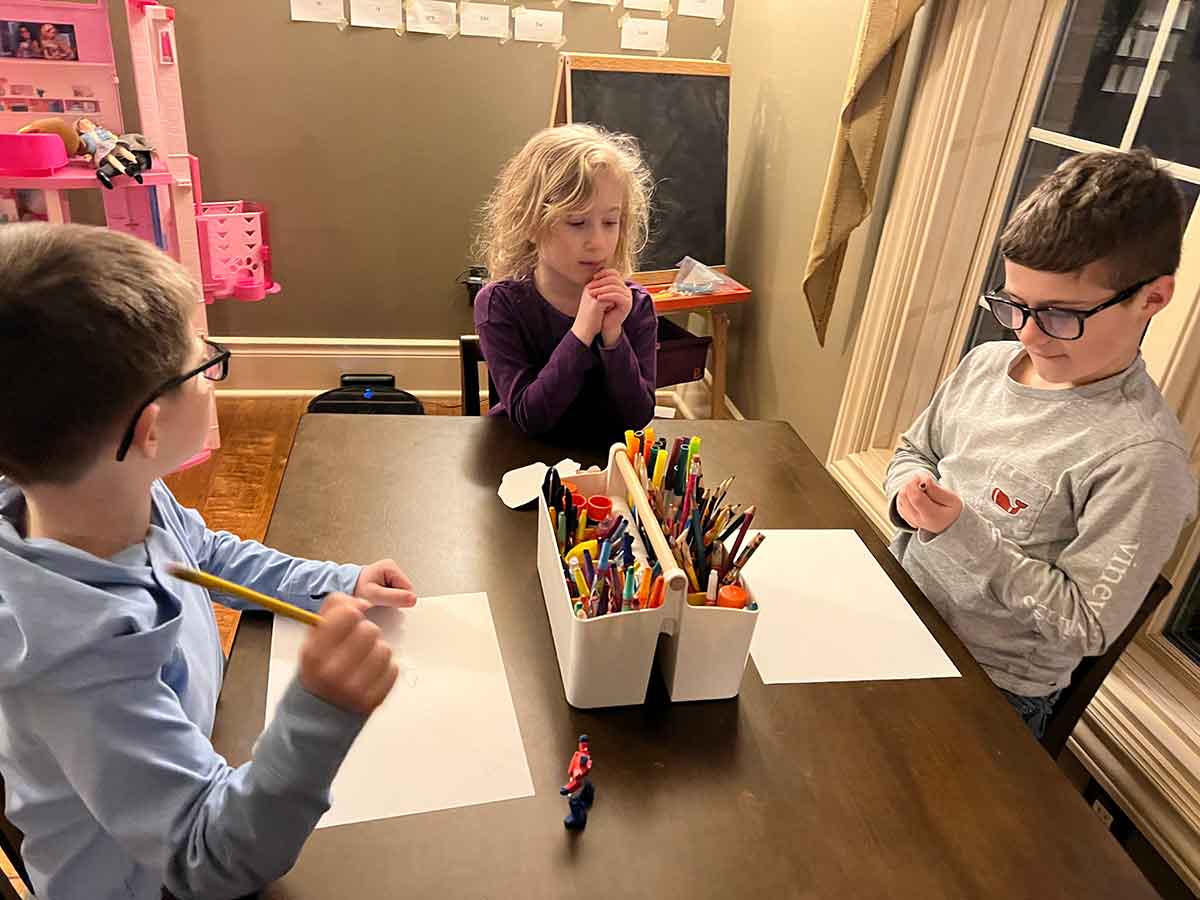 Two boys and a girl sitting at a table with art supplies and pieces of paper.