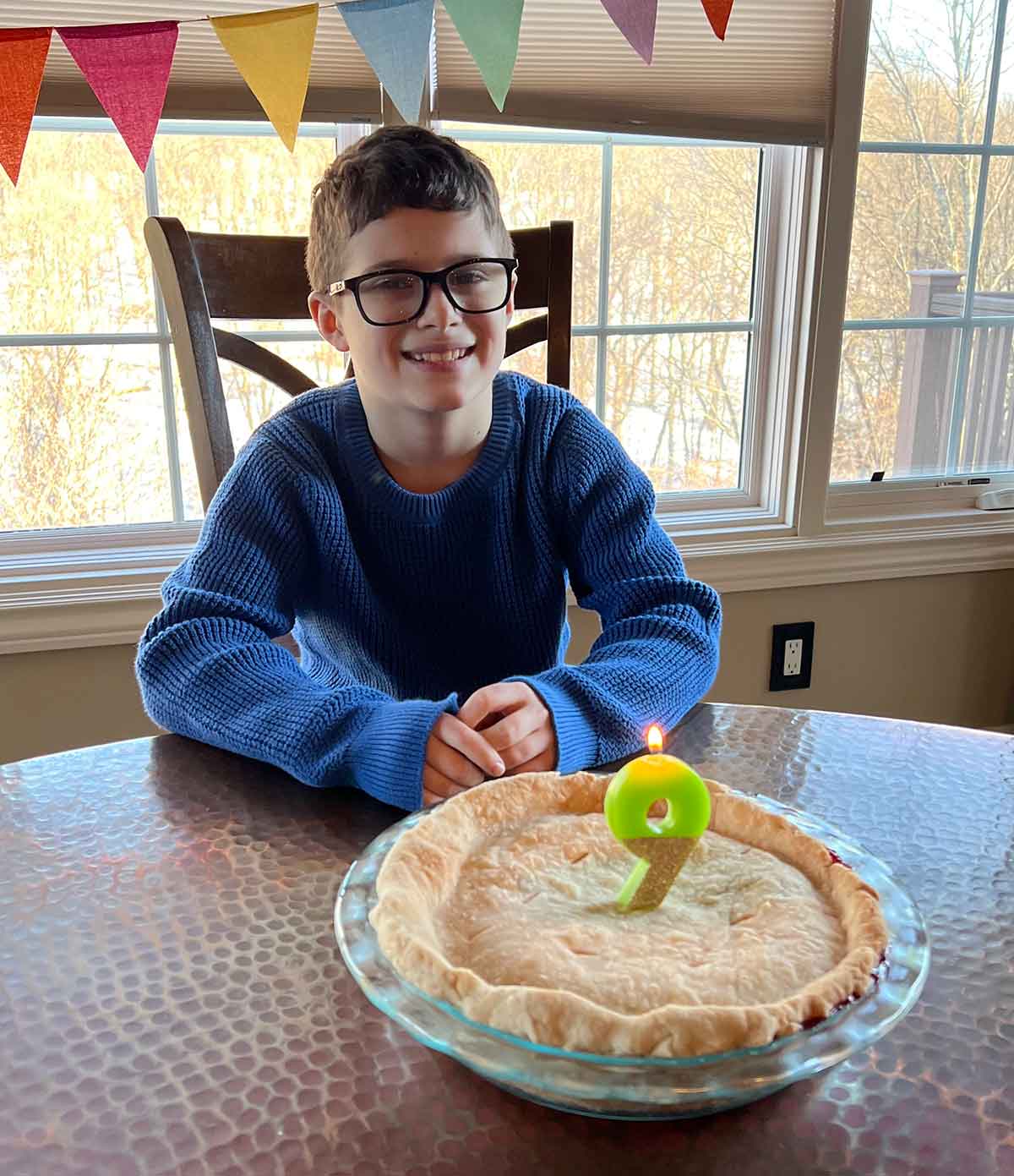 Boy in a blue sweater and black glasses sitting in front of a pie with a number nine candle in the middle of it.