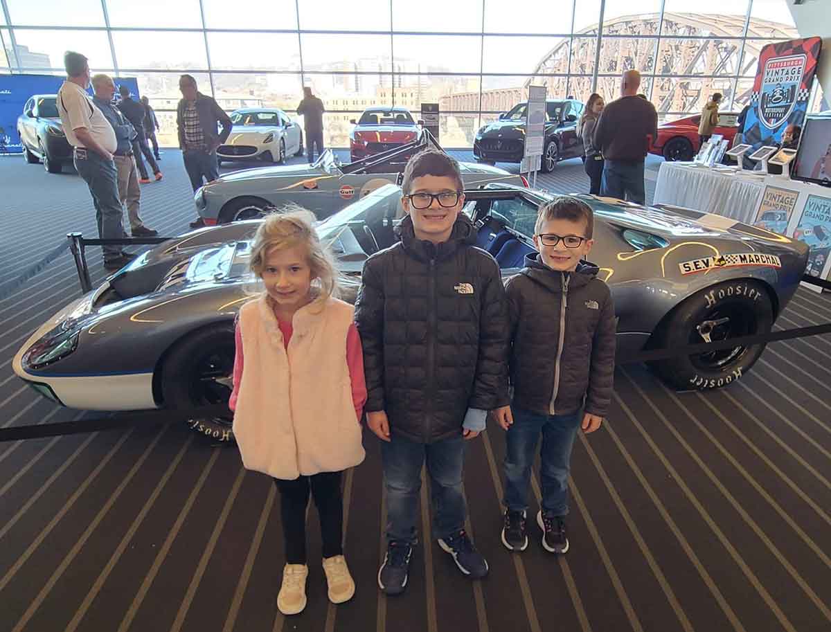 A girl and two boys standing in front of a race car at an auto show.