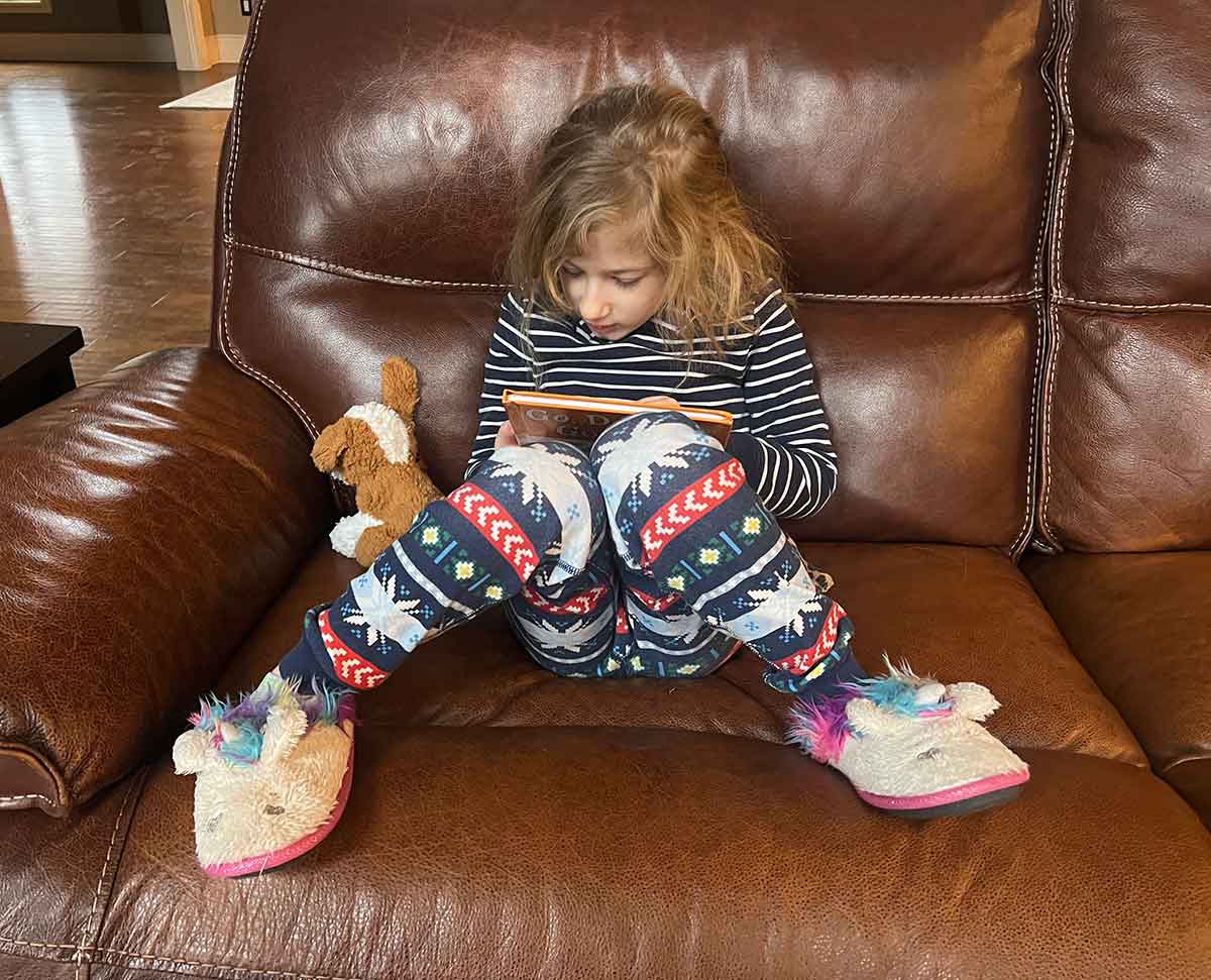 Little girl sitting on the couch in pajamas and slippers, reading a book with a stuffed animal next to her.