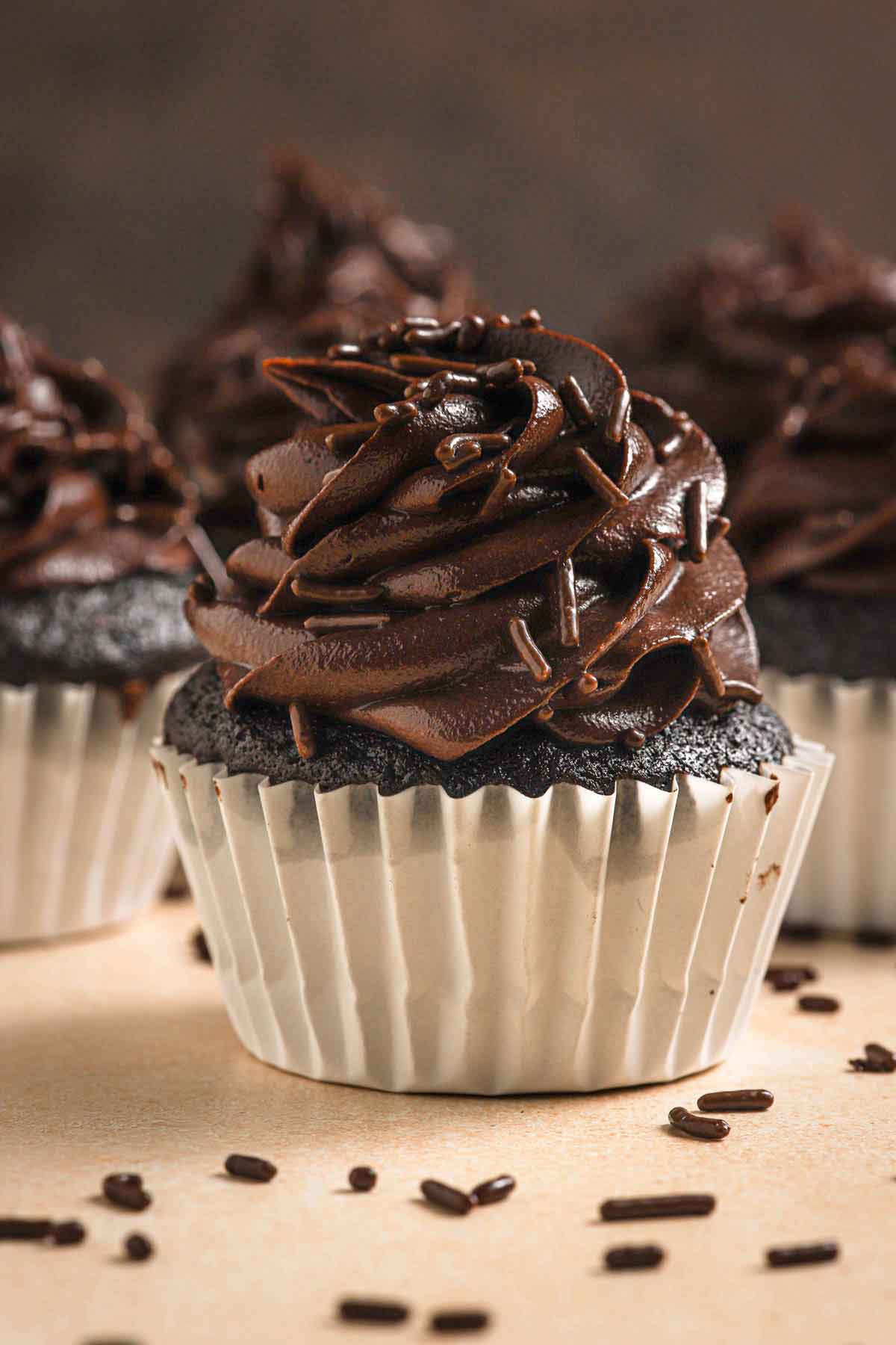 Head-on close-up photo of a chocolate cupcake in a white paper liner with chocolate frosting and chocolate sprinkles.