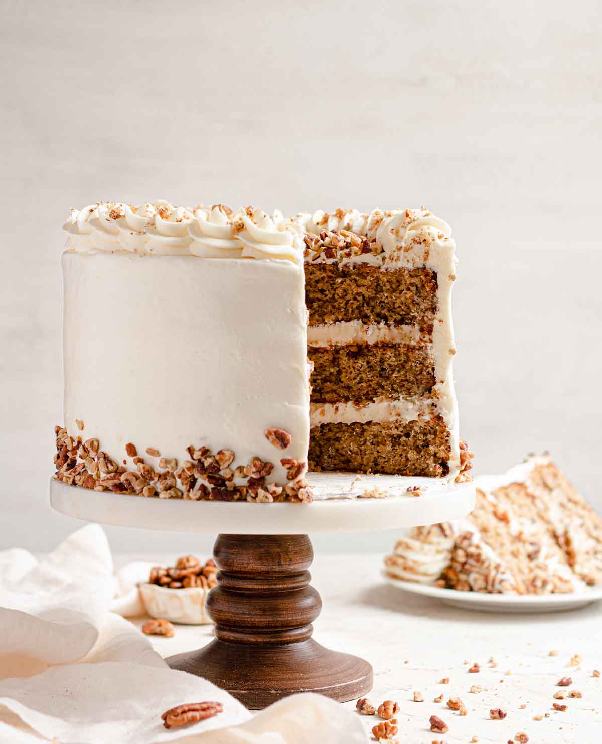 Side view of three-layer hummingbird cake with a slice removed, filling and frosted with cream cheese frosting and garnished with chopped pecans.