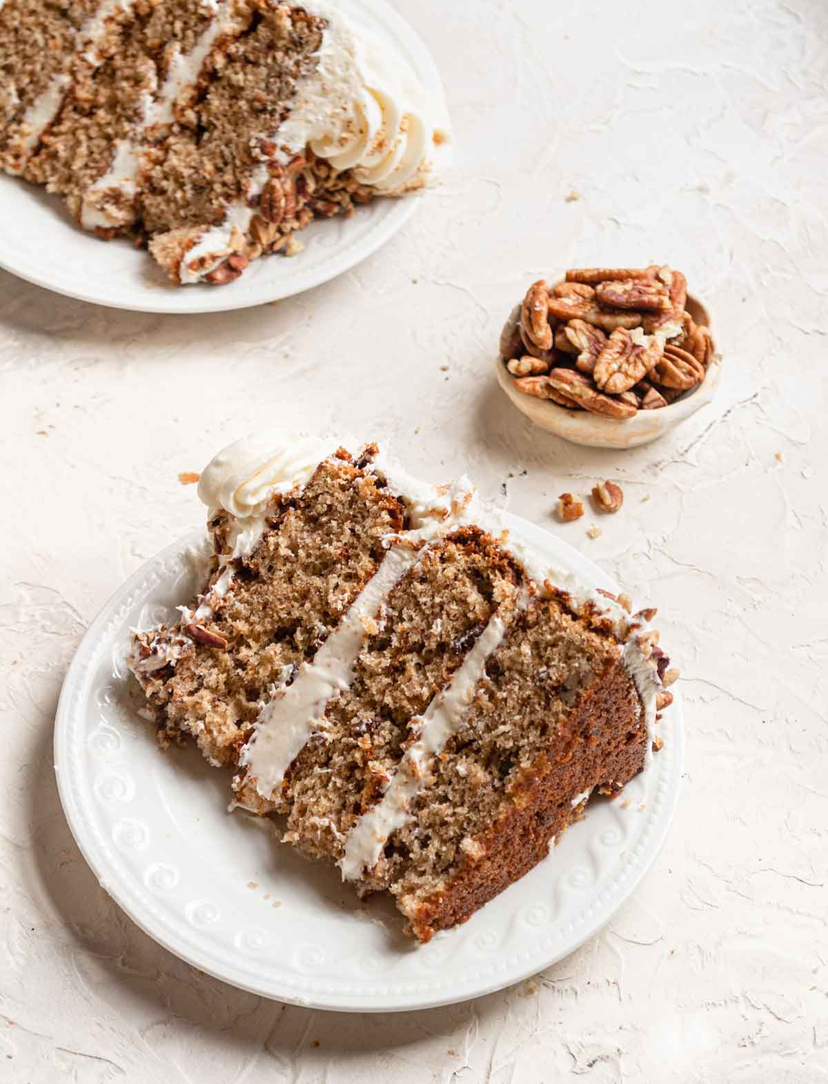 Slice of hummingbird cake on a white plate with a small bowl of pecans in the background.