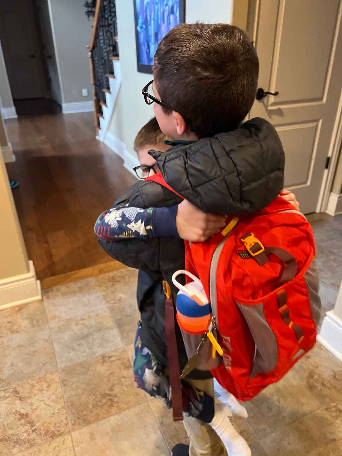 A boy in a jacket and backpack hugging and holding a smaller boy in pajamas.
