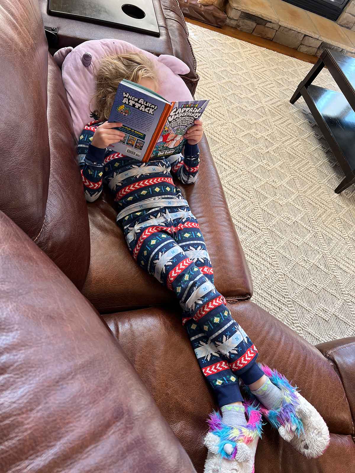 Little girl laying on a couch with a squishmallow and reading a Captain Underpants book.