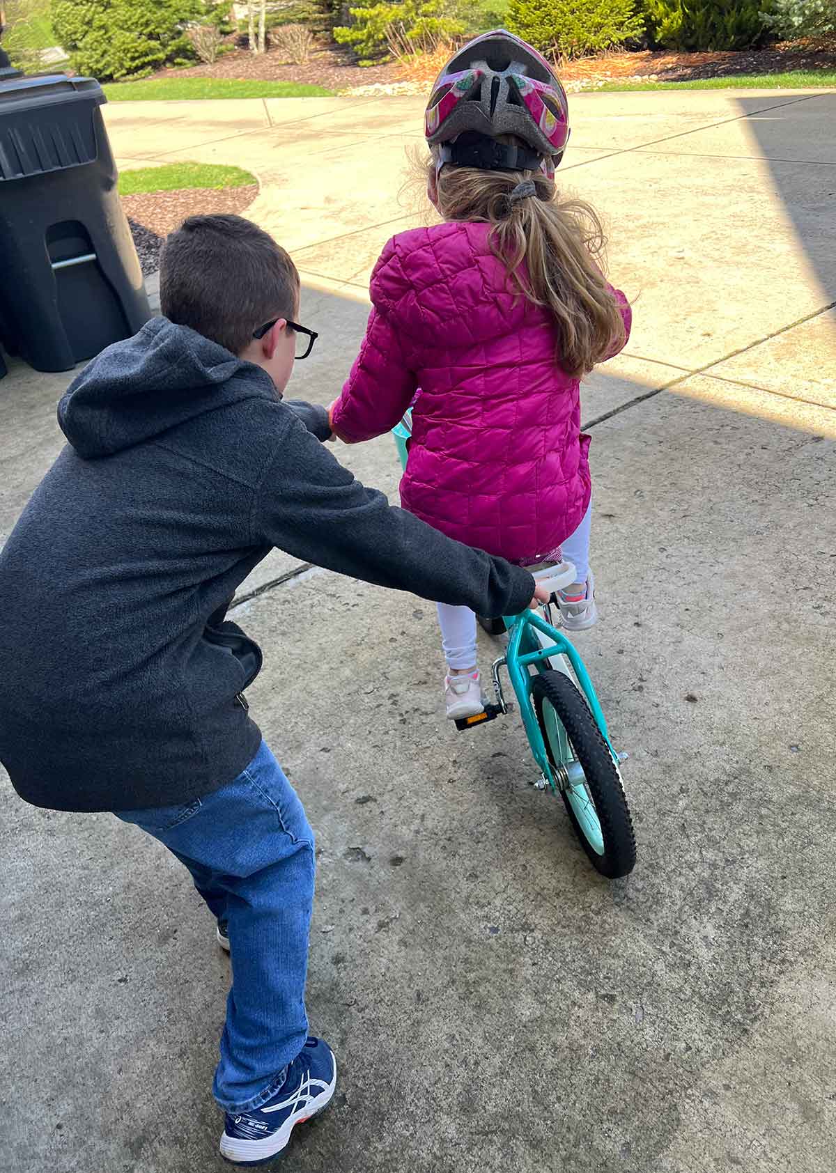 A boy helping a girl in a pink jacket on a bike in a driveway.