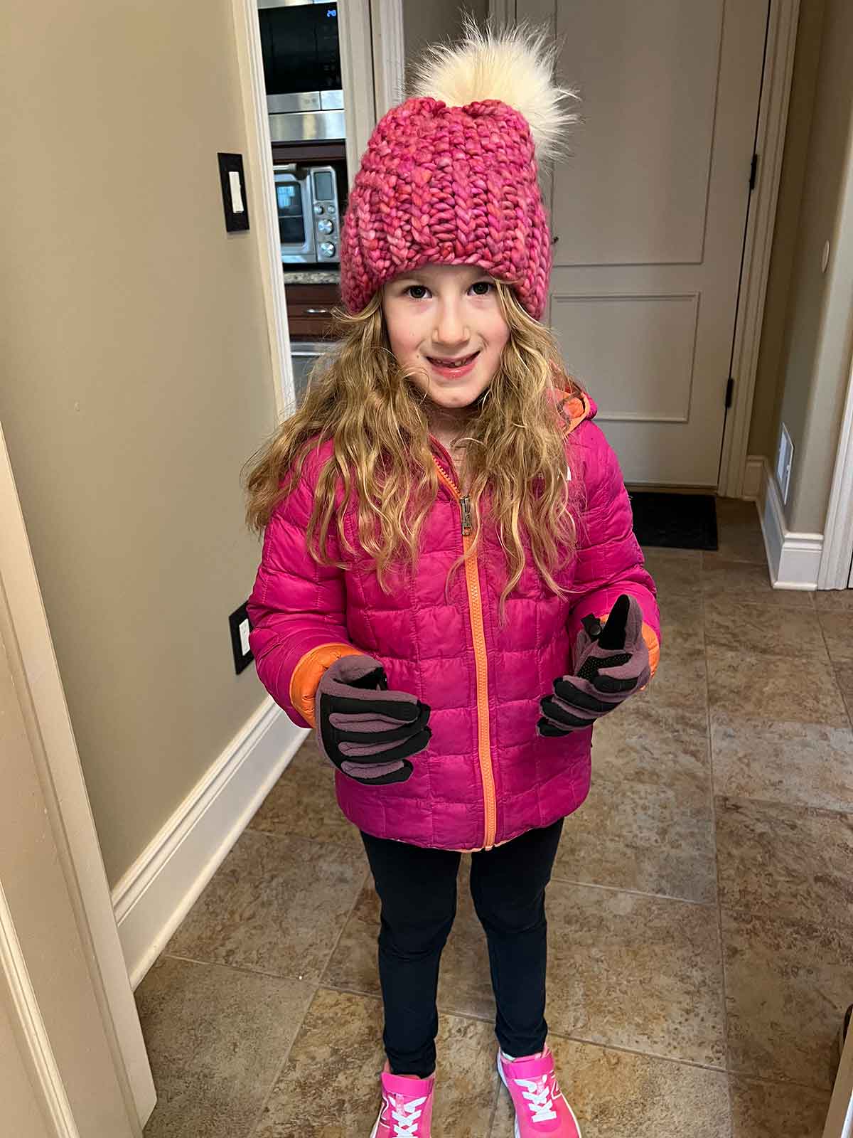 Little girl with blond curly hair in a pink winter jackets, pink hat, and gloves.