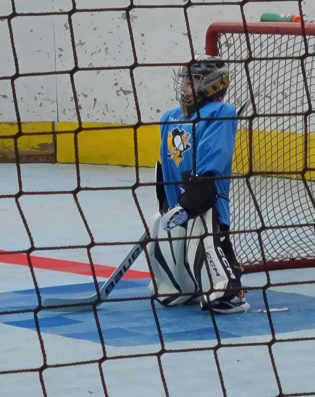 Little boy in a blue jersey and goalie equipment standing in front of a hockey net on a dek surface.