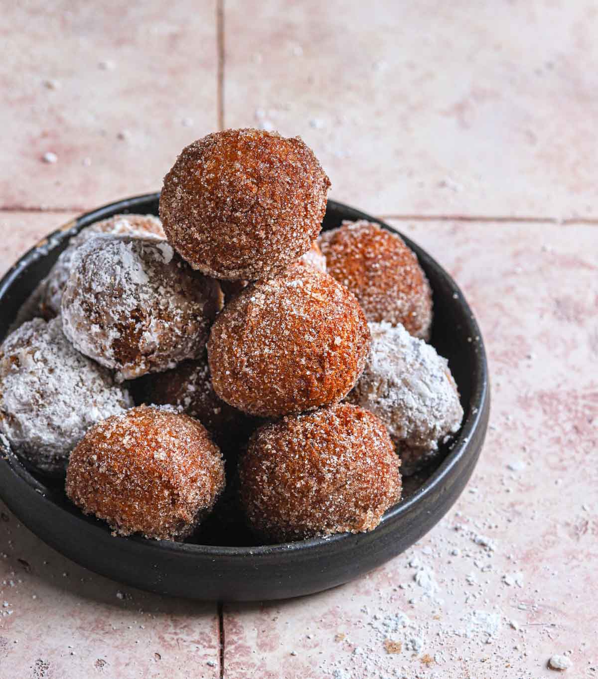 Fried zeppole tossed in powdered sugar and cinnamon sugar stacked in a low black bowl.