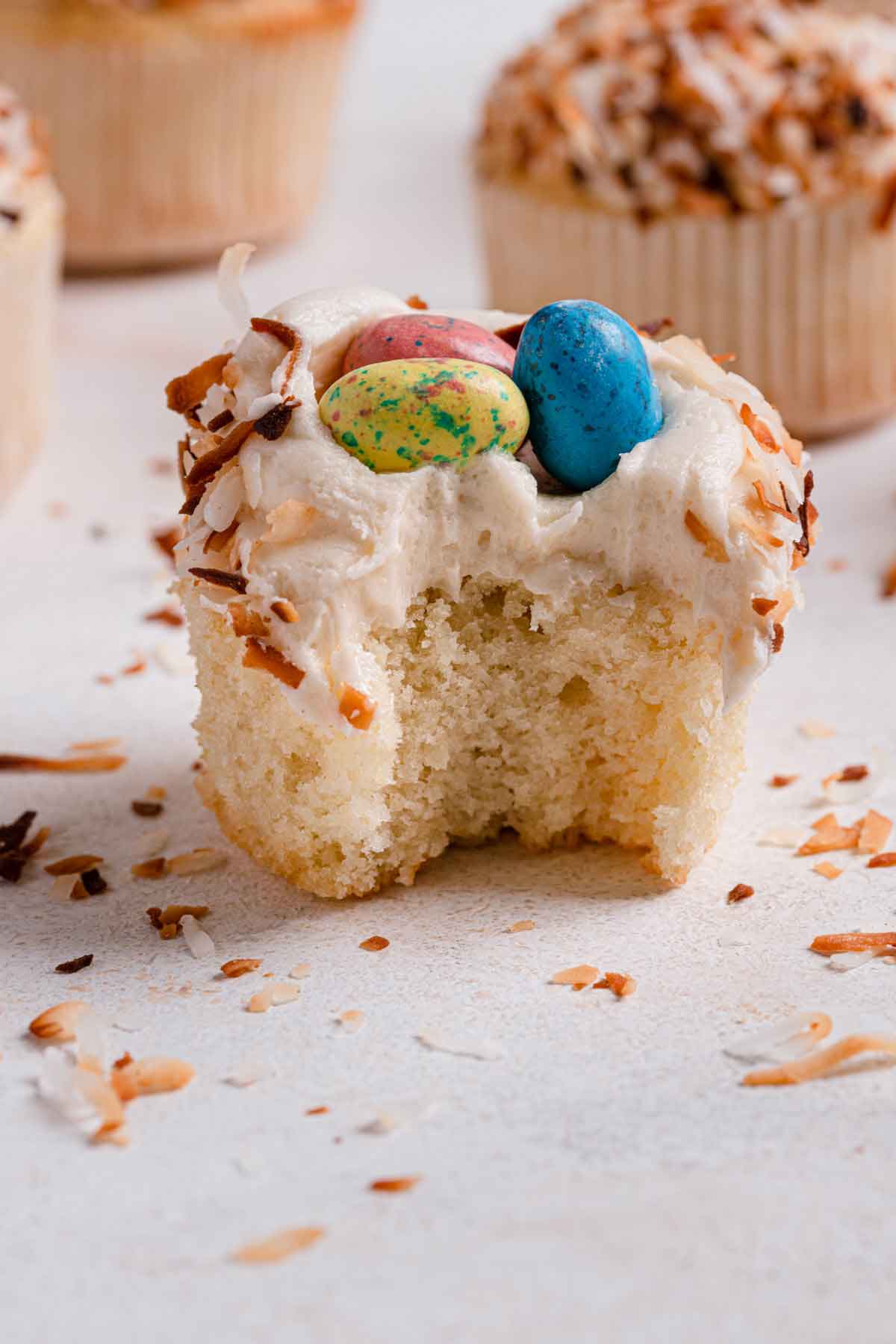 Single coconut cupcake with coconut frosting, topped with toasted coconut and malted eggs, with a bite taken out.