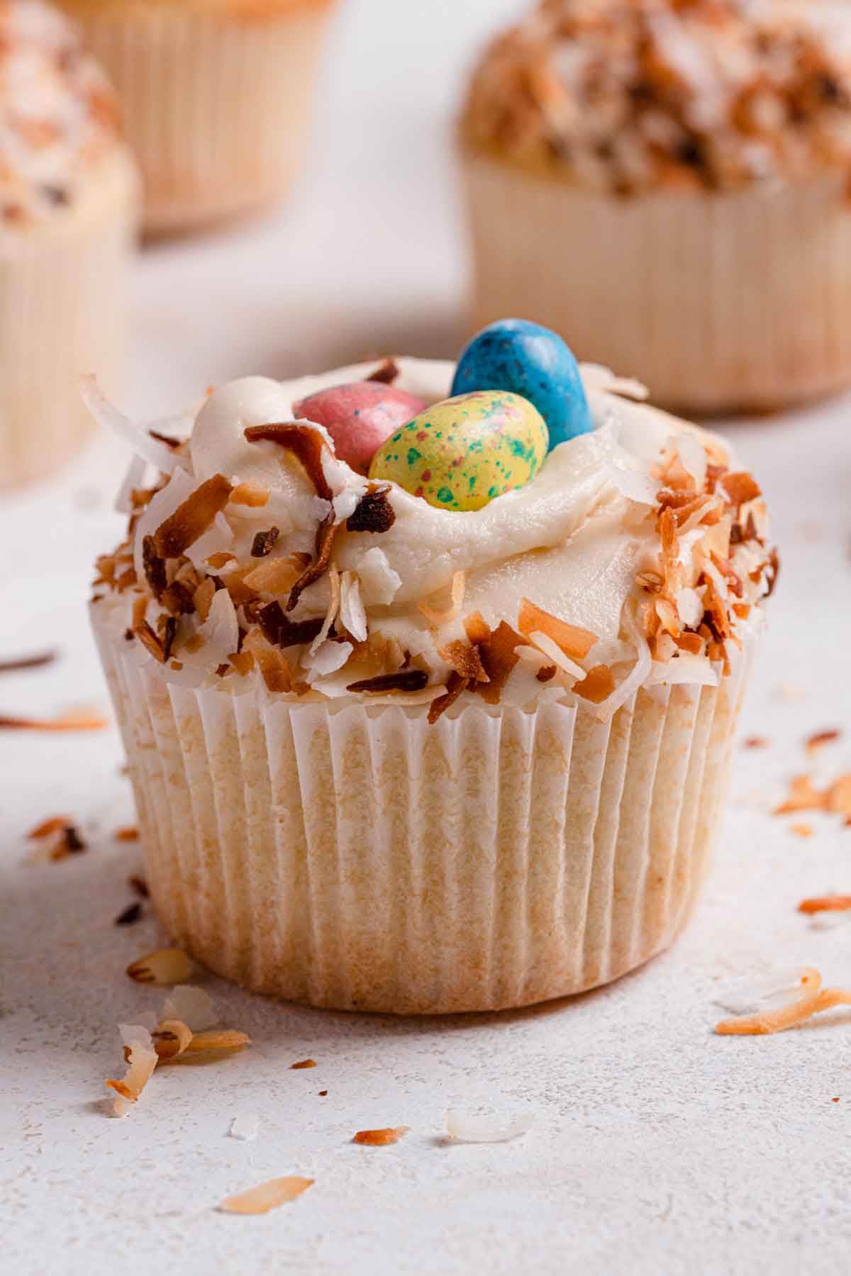 A single coconut cupcake in a paper wrapper, topped with frosting, rolled in toasted coconut flakes and topped with Whopper eggs.