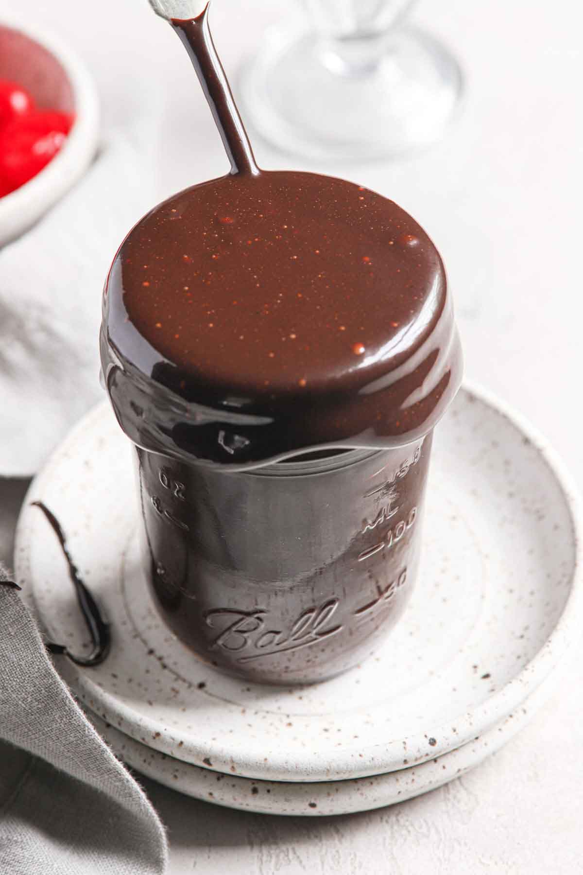 Glass jar overflowing with hot fudge sauce, sitting on top of two stacked white speckled plates.