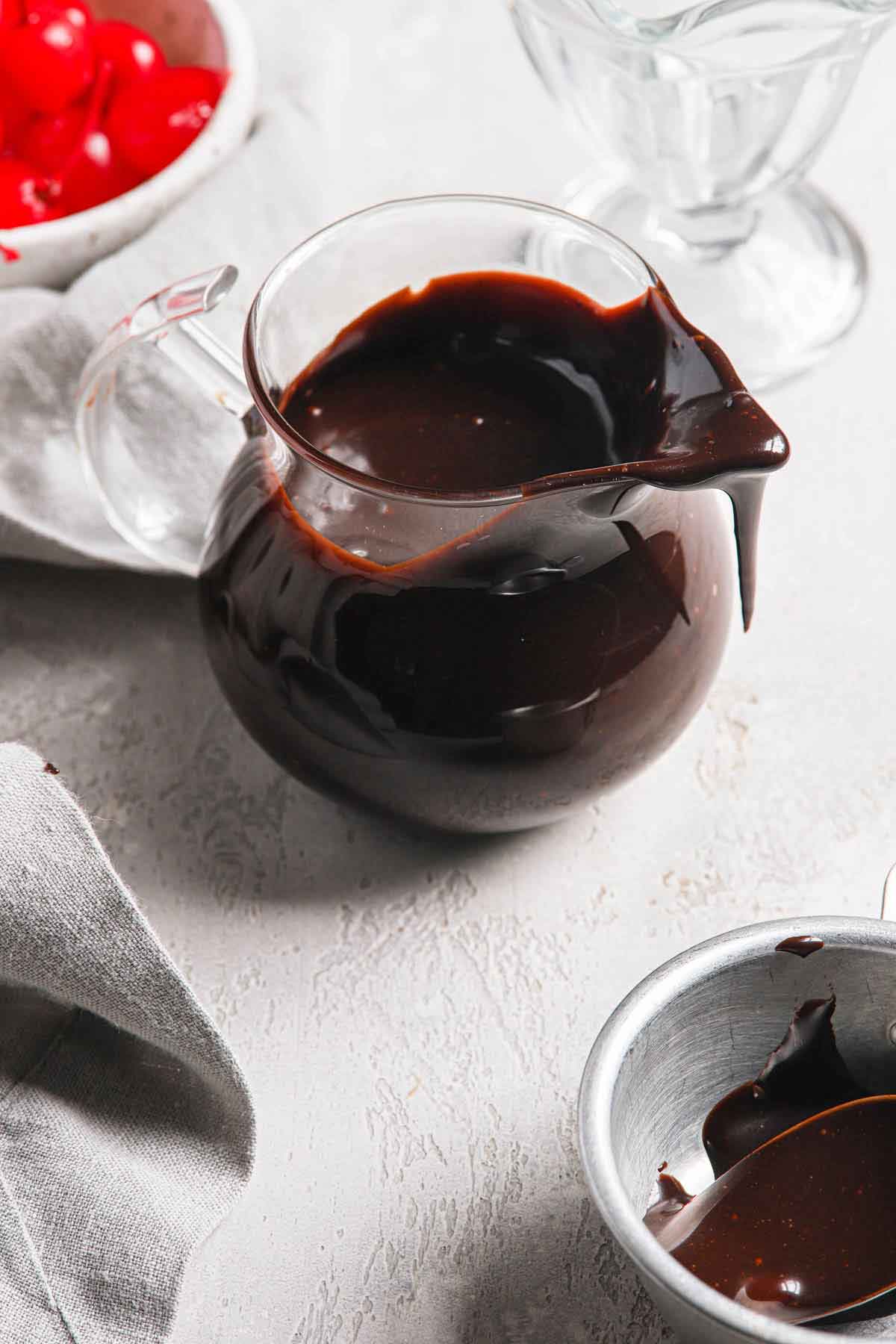 Glass pitcher full of hot fudge sauce with a bit of sauce dripping off the lip of the pitcher.