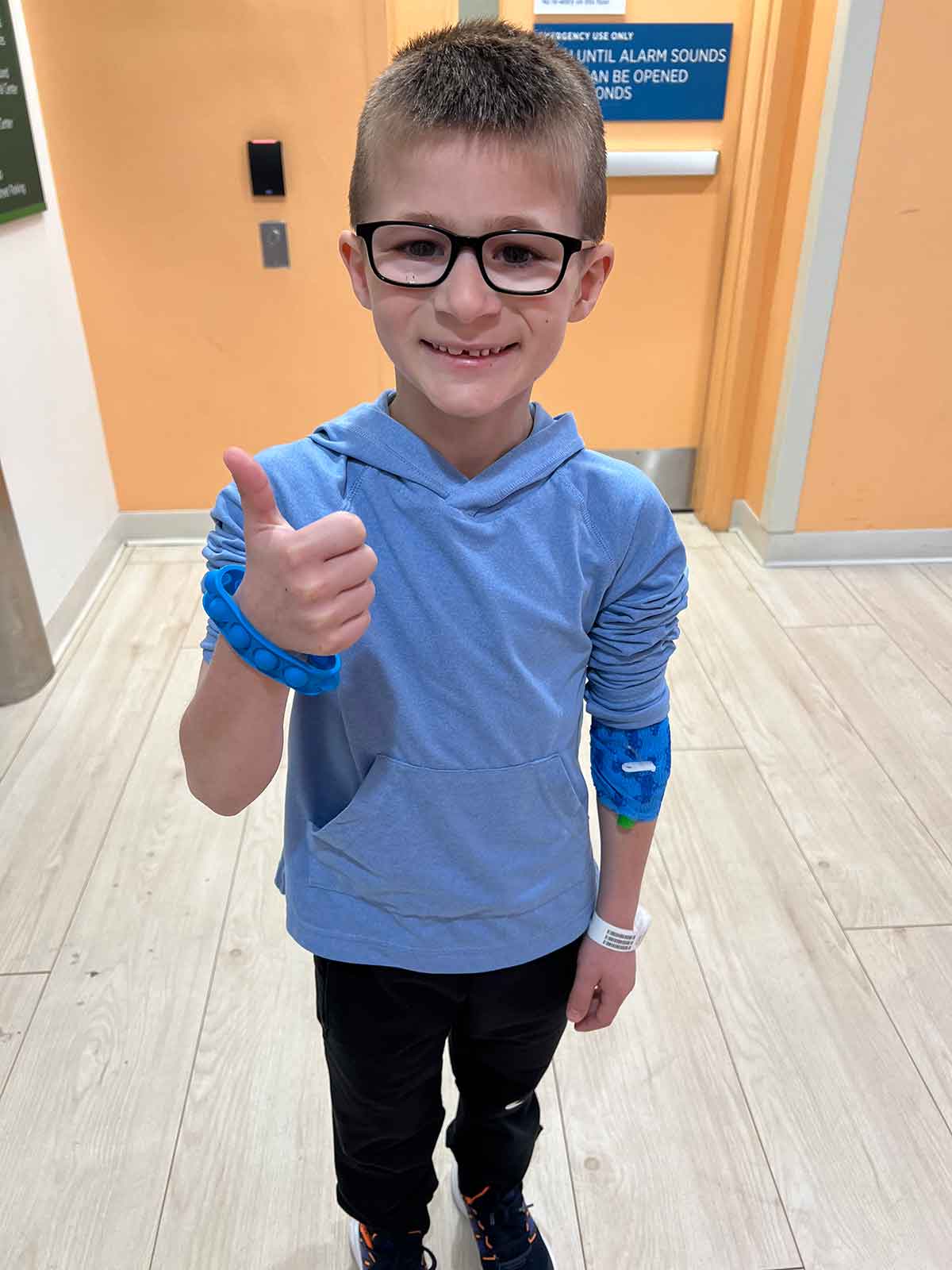 Little boy with black glasses and a blue hoodie, smiling and giving a thumbs up.