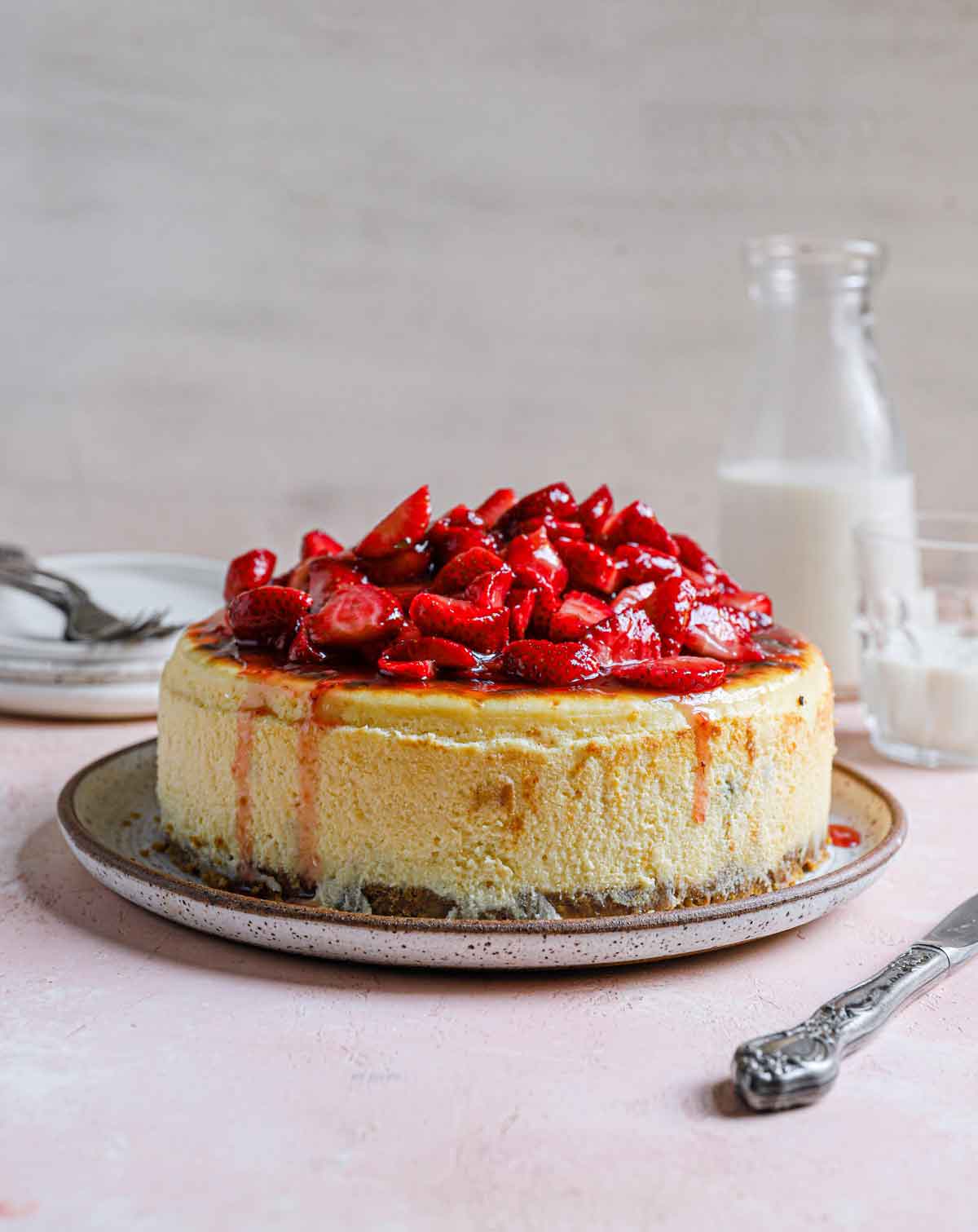 Straight on view of New York cheesecake with fresh strawberry topping with a glass of milk in the background.