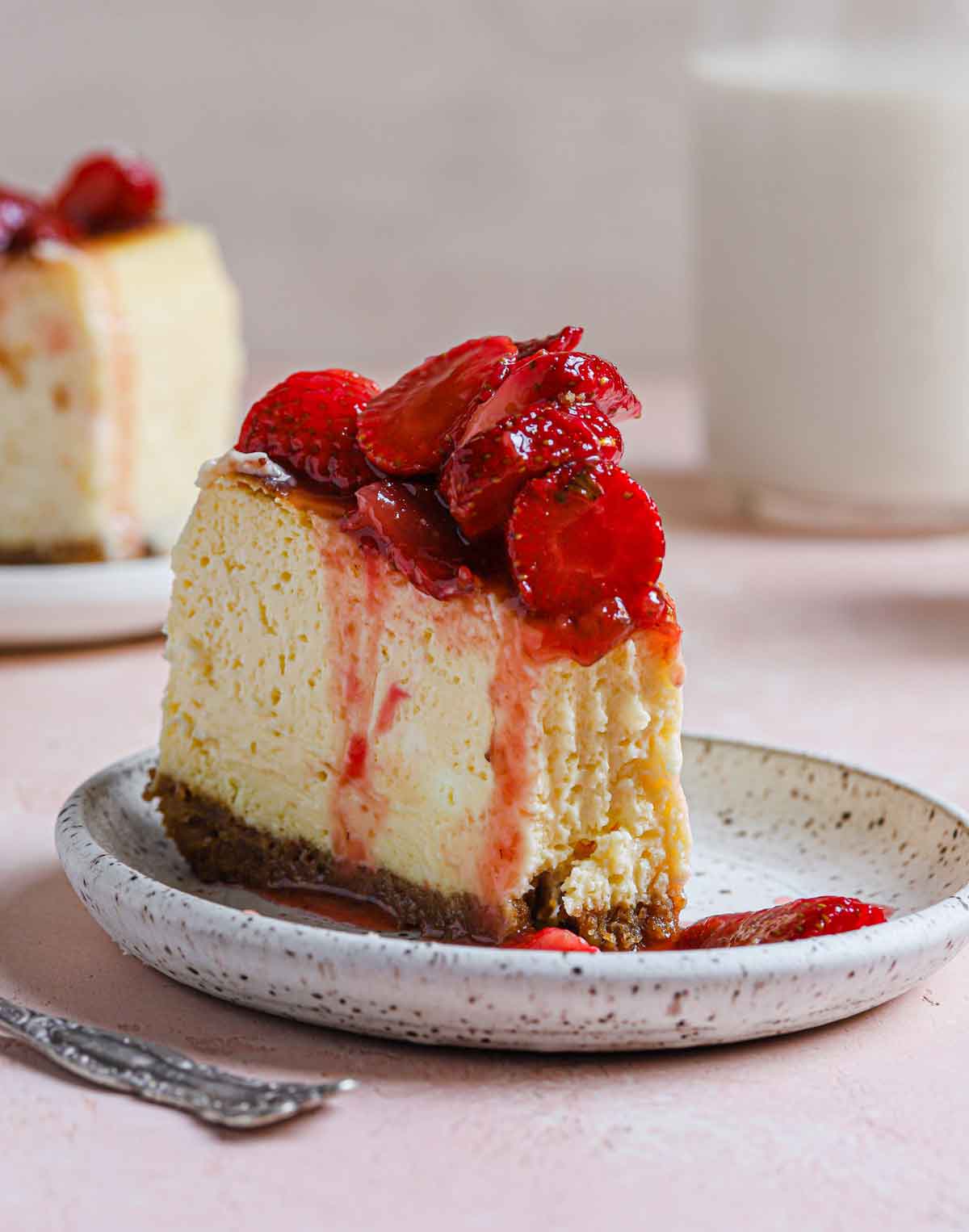 Slice of New York cheesecake with strawberry topping on a white and black speckled plate.