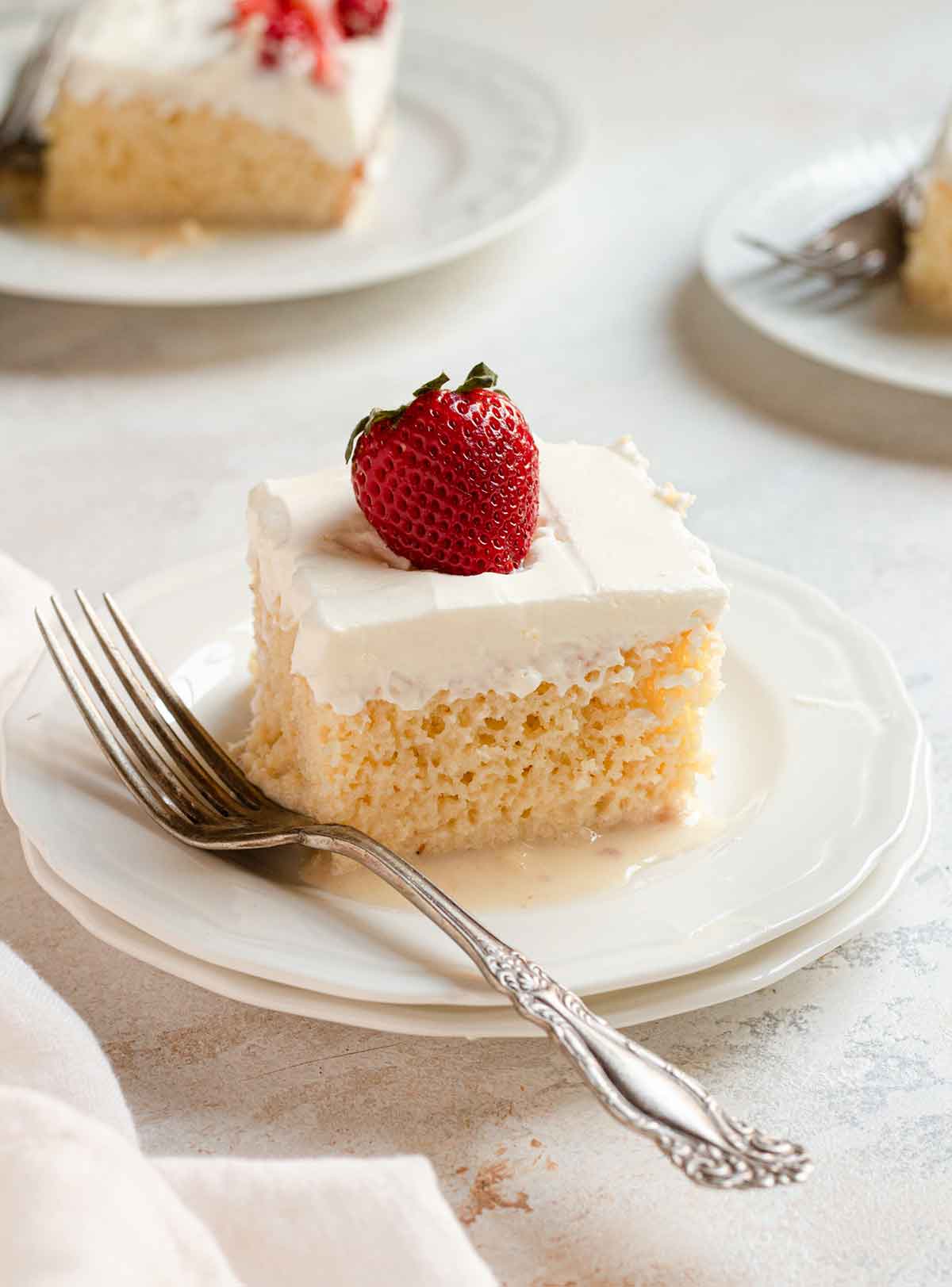 Slice of tres leches cake with a strawberry on top, sitting on a white plate with a fork to the side.