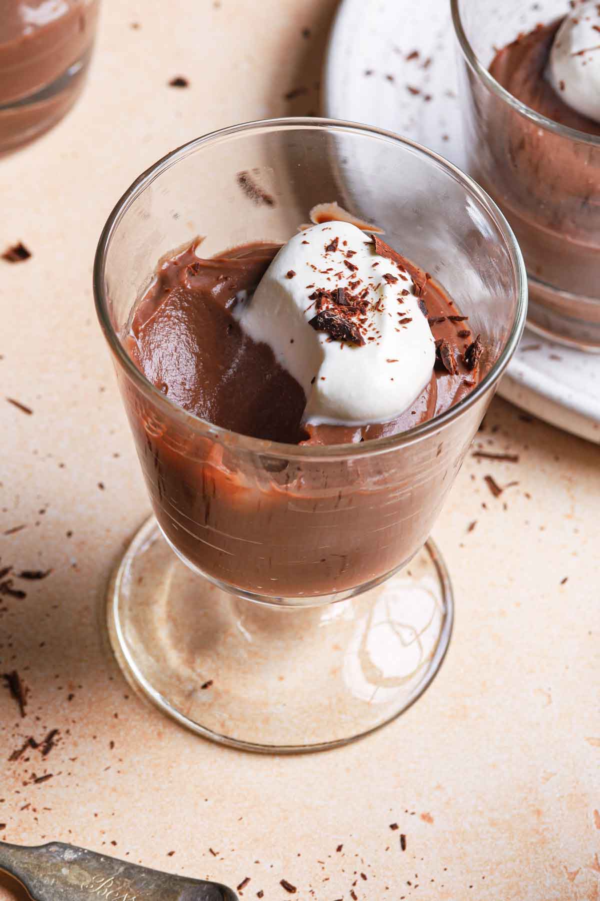 Close up photo of a glass serving cup with chocolate pudding topped with whipped cream and chocolate shavings, with a spoonful taken out.