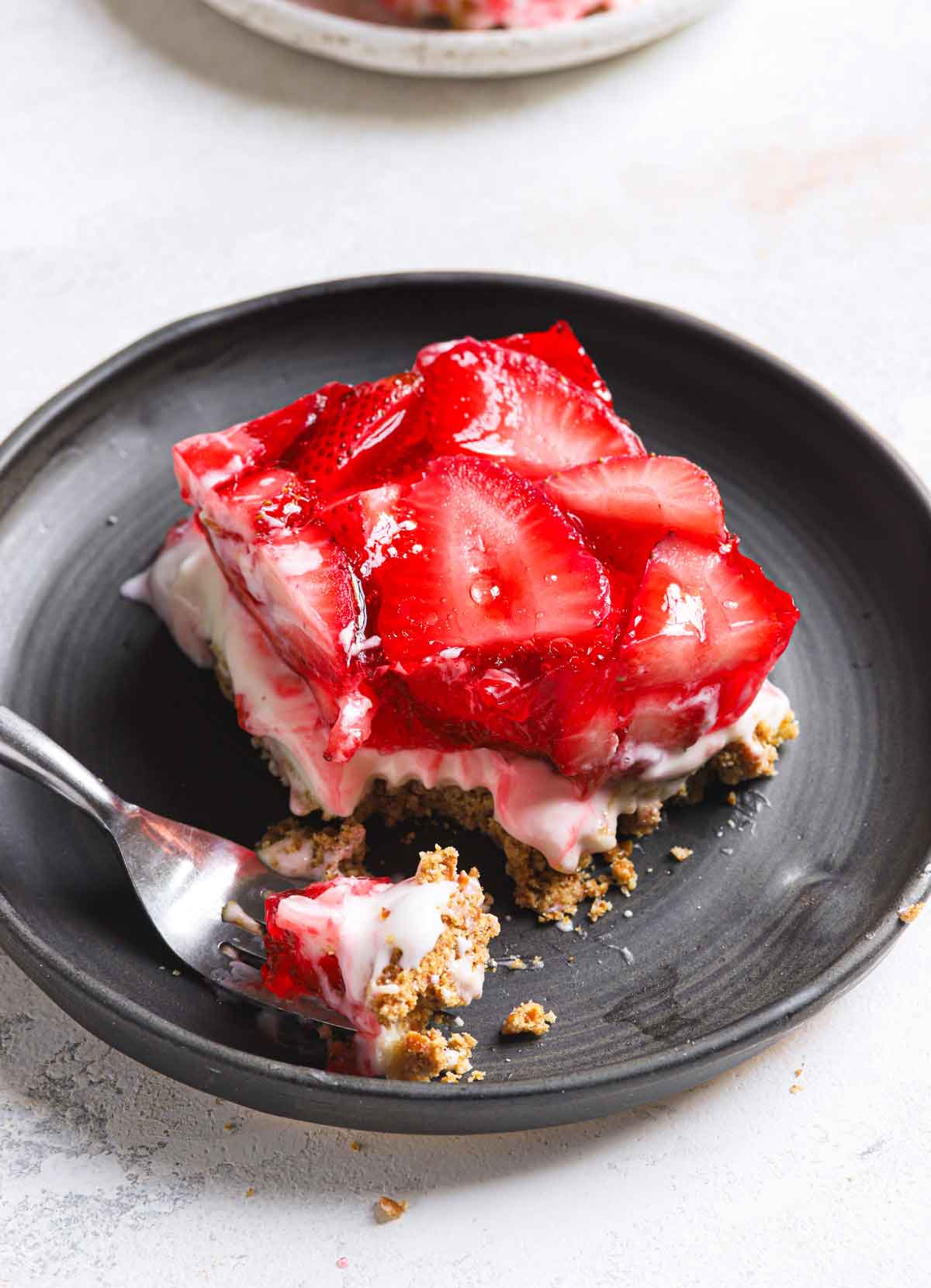 A square of strawberry pretzel salad with a forkful removed on a dark gray plate.