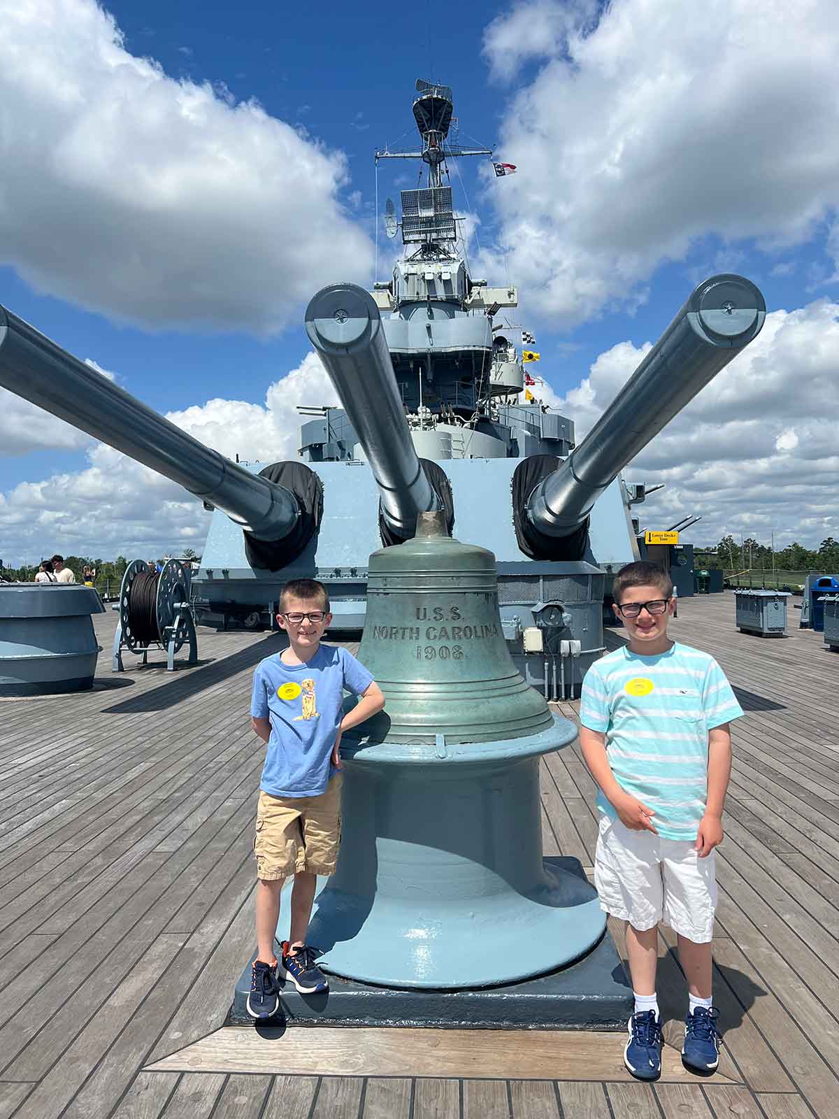 Two boys standing on either side of a bell labeled "U.S.S. North Carolina 1908" on the top deck of a battleship.