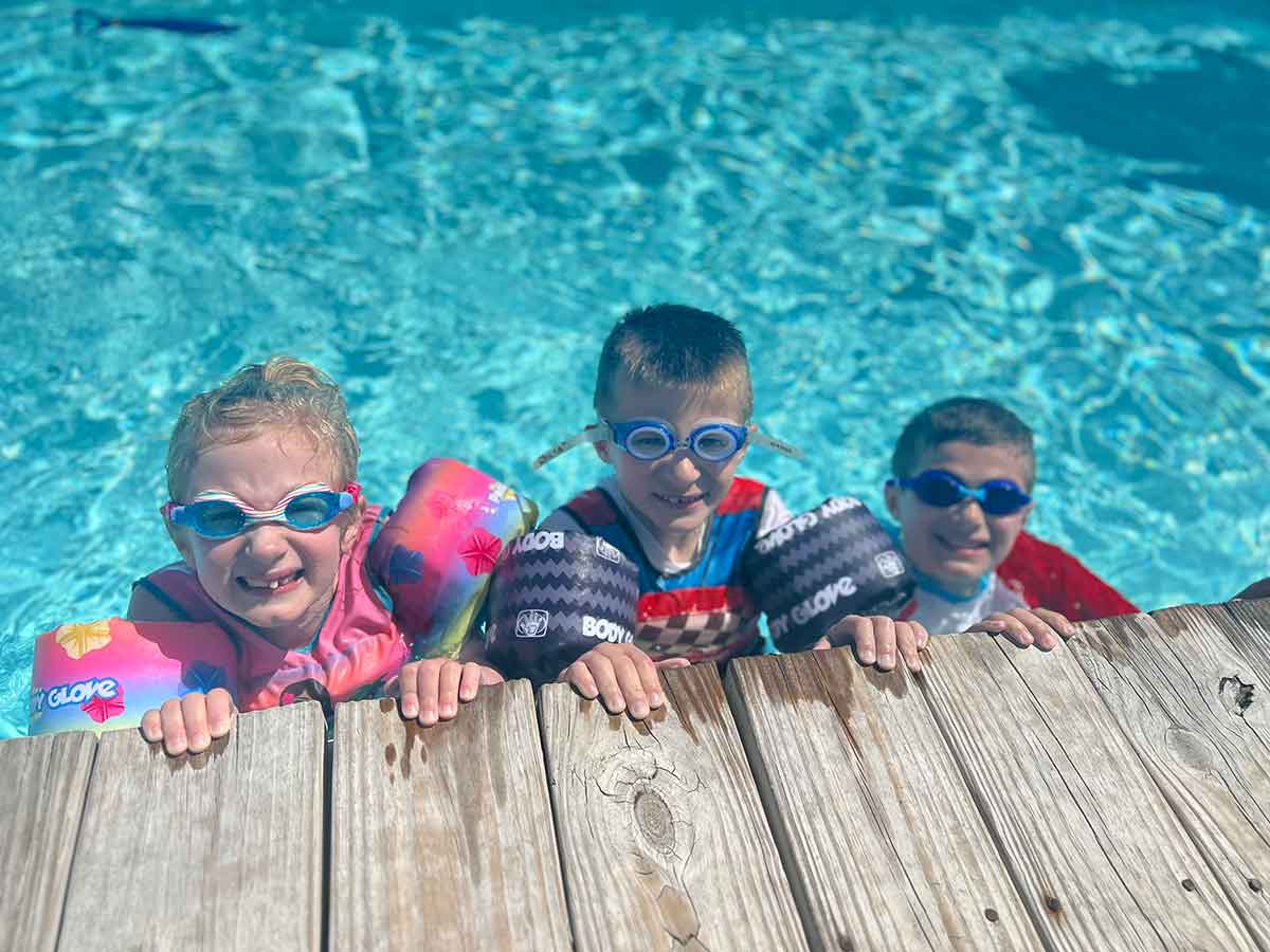 Three kids in a pool, hanging onto the side, smiling.