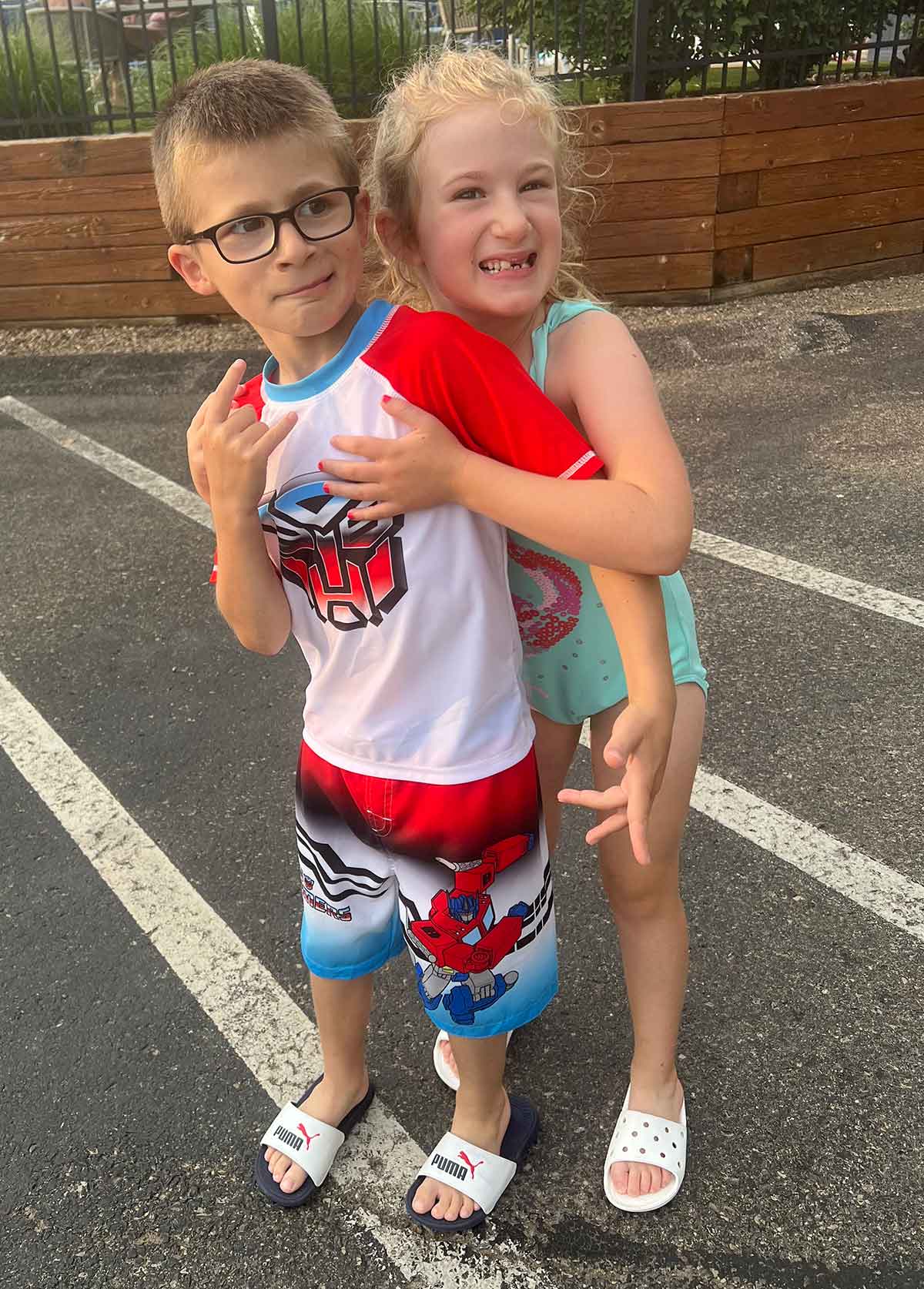 Little girl hugging a boy from behind, both in swimsuits in a parking lot.