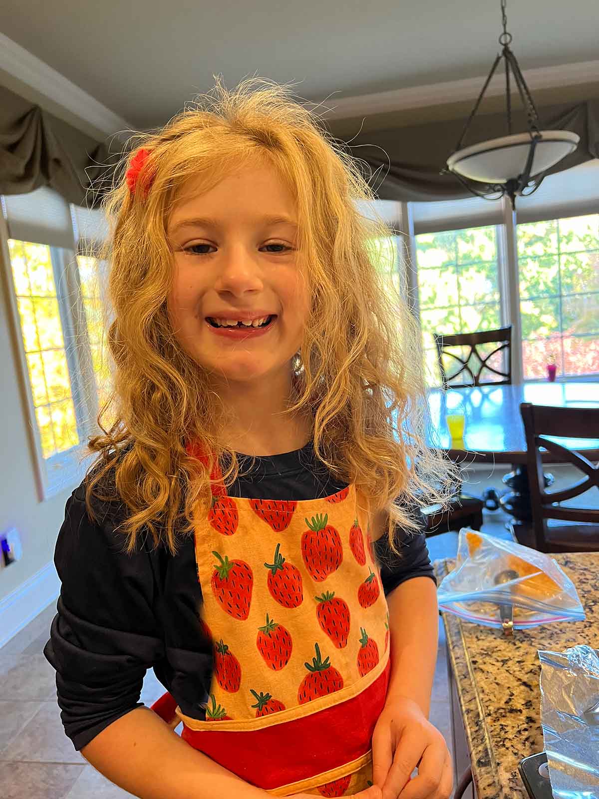 Little girl in a strawberry apron standing at a kitchen counter.