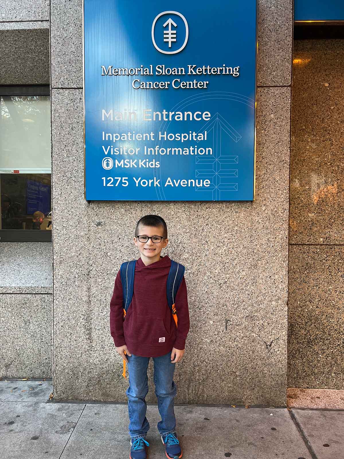 Little boy with a backpack standing below a sign at the entrance of Memorial Sloan Kettering Cancer Center.