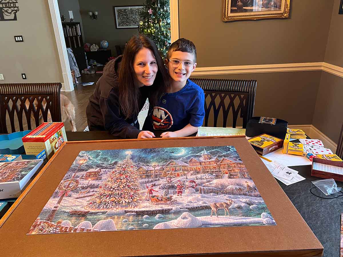 A mom and a boy standing together behind a completed jigsaw puzzle on a dining room table.