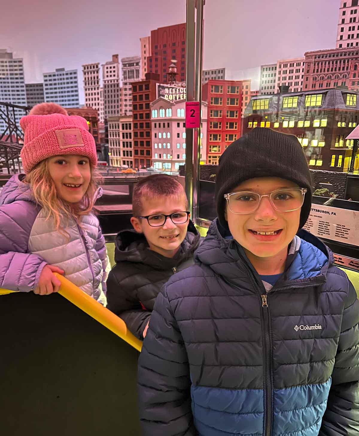 Two boys and a girl in winter coats standing in front of a model train display.