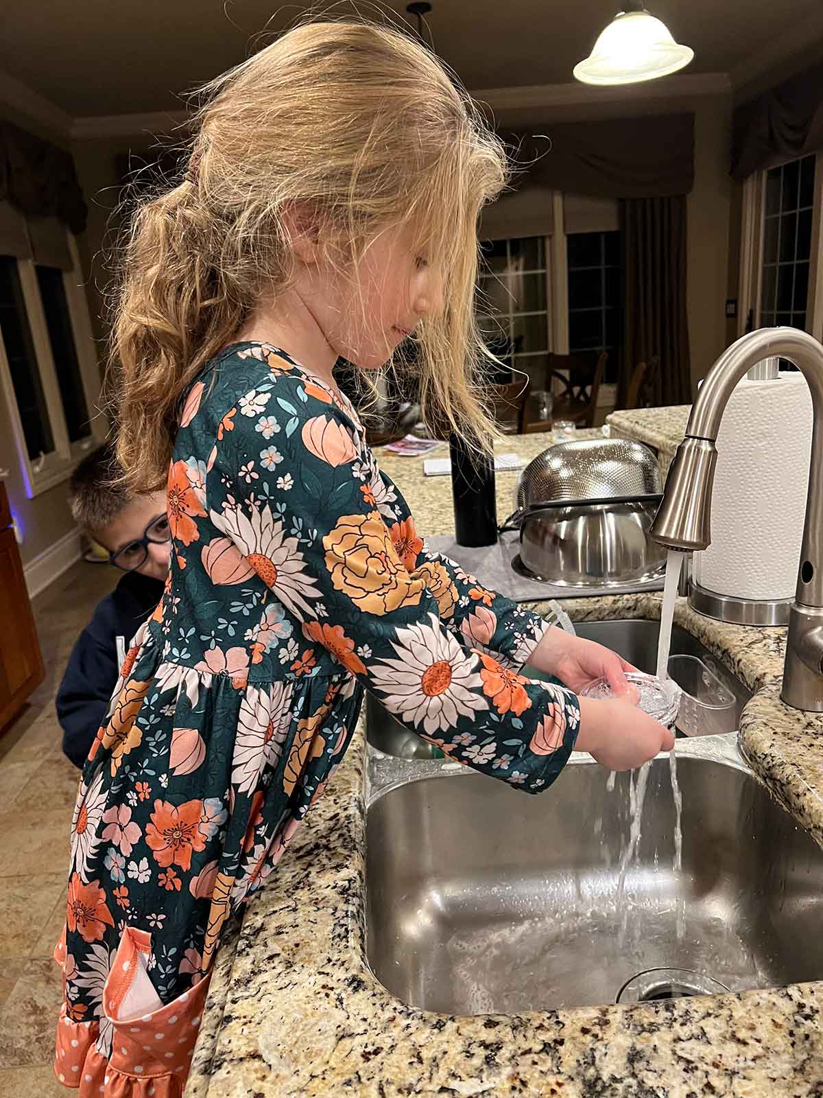 Little girl in a floral dress washing dishes at a kitchen sink.