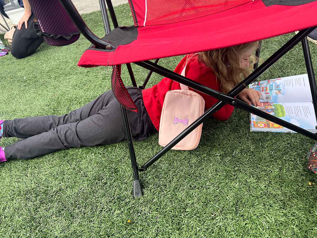 Little girl lying on a field unde a red camping chair reading a book.